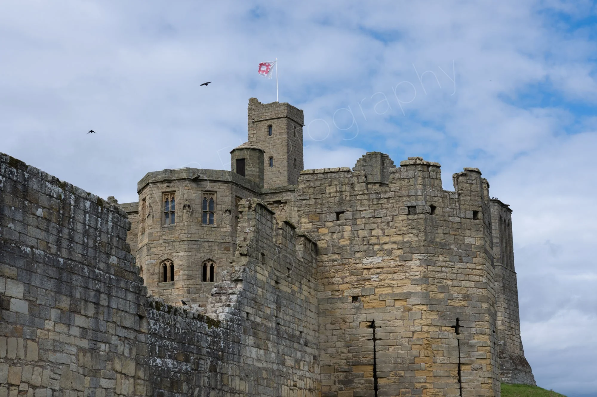 Part of Warkworth Castle, Northumberland with a damaged battlements wall creating a view to the Great Tower, cloudy blue sky background.