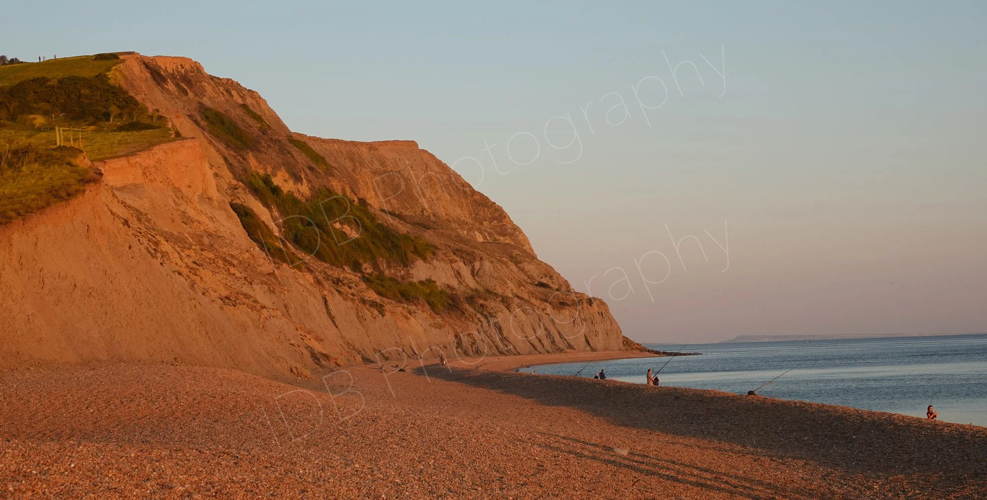 Seatown beach on the beautiful Jurassic Coast, Dorset nearing sunset, the cliffs and shingle beach appears extra red, anglers fish below a tranquil hazy blue sky.