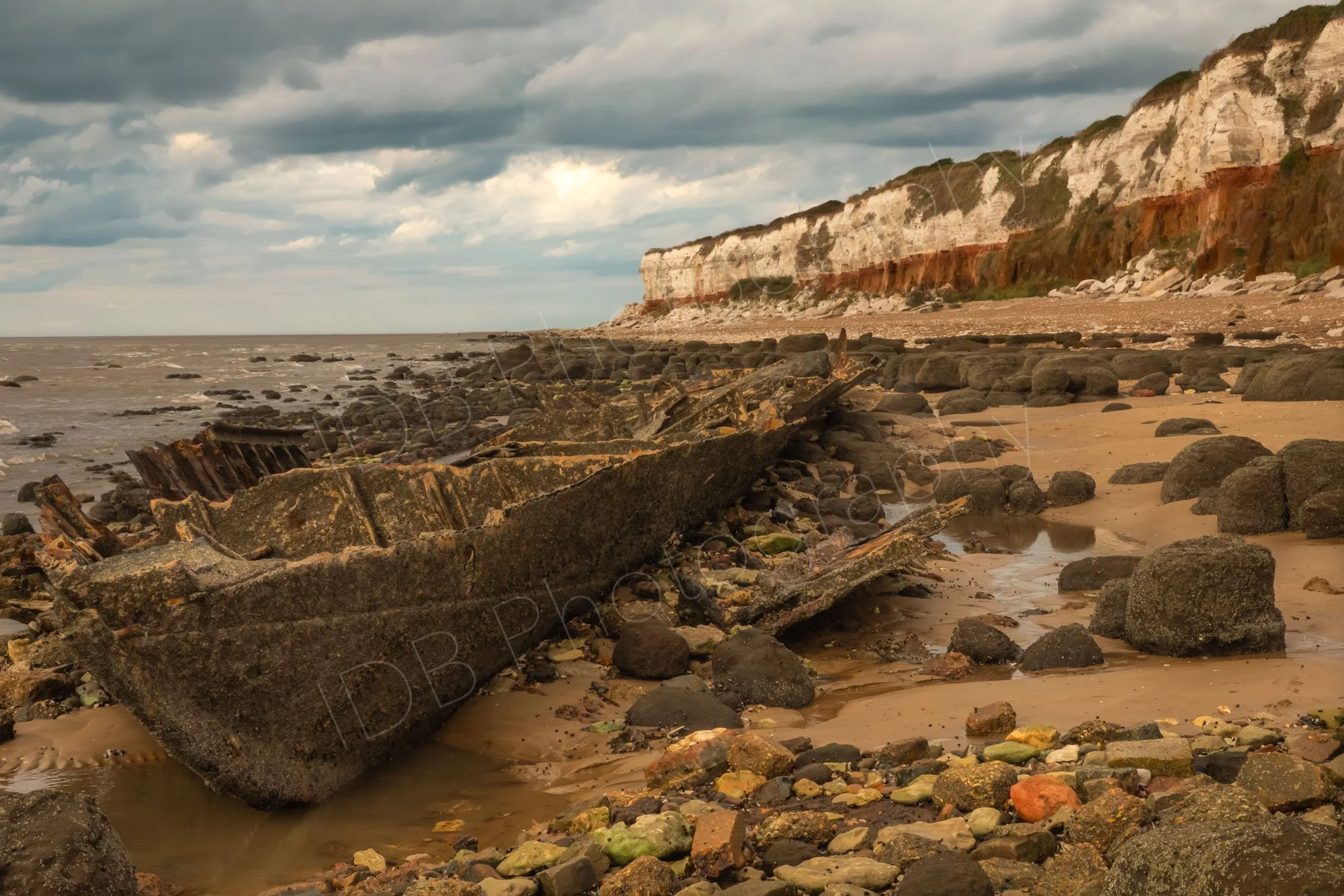 A view along an old boat wreck at Hunstanton Beach, Norfolk,  coloured stones in the foreground and stunning bicolour cliffs behind under a stormy sky.