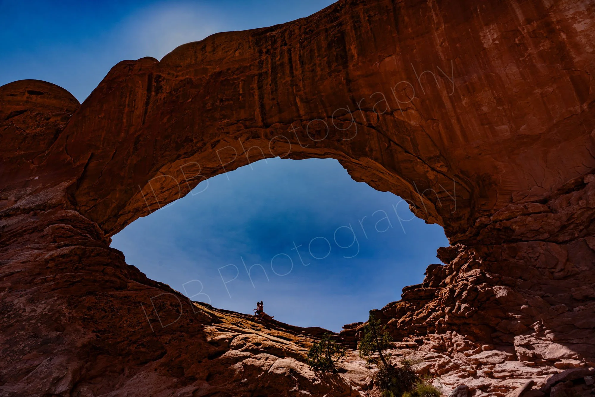 North Window Arch in Arches National Park, near Moab, Utah with 2 people sitting on the lower rim, variable blue sky background.