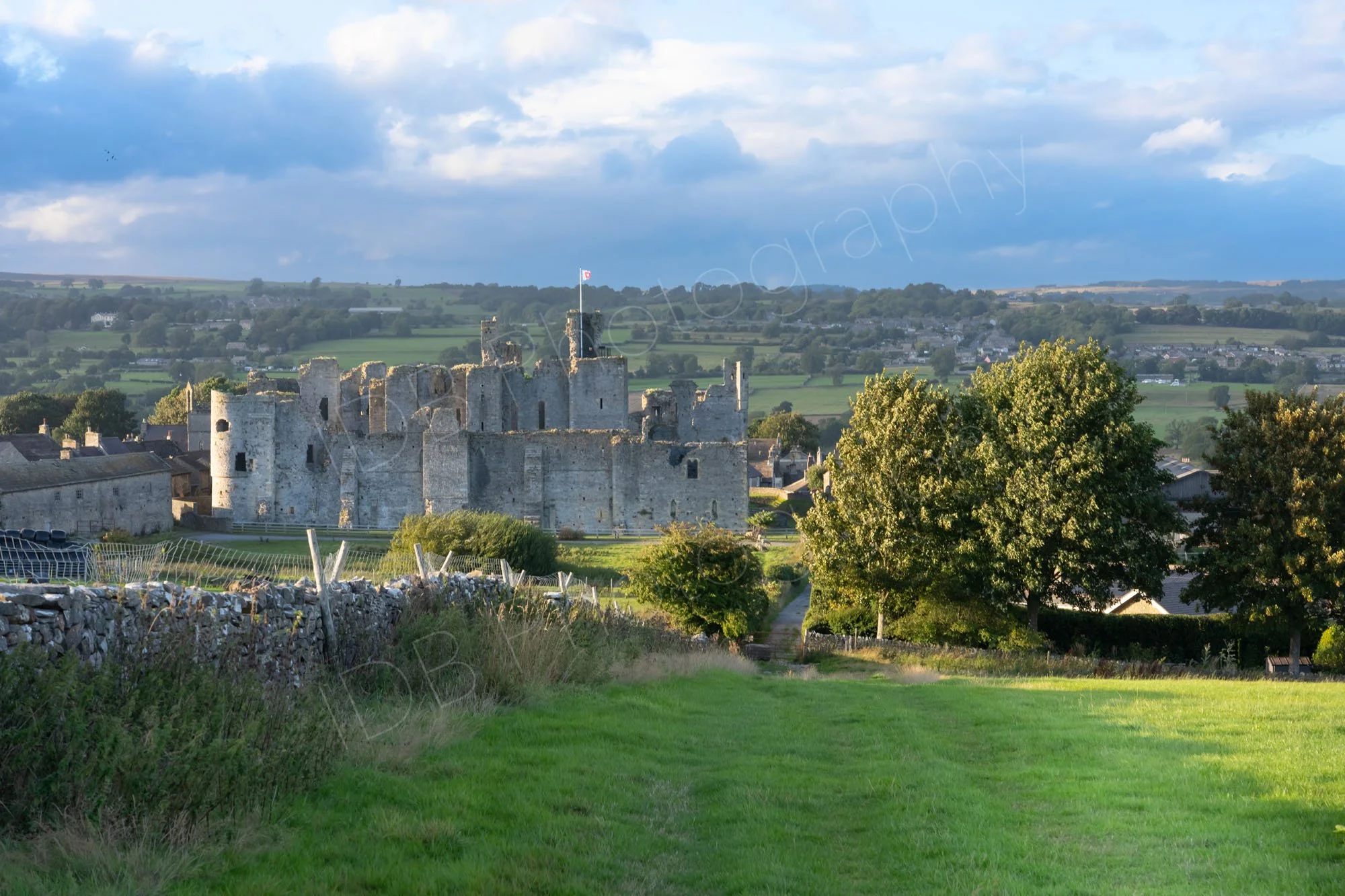 A view down a dry stone wall of Middleham Castle, Yorkshire, with trees, distant landscape and cloudy blue sky.