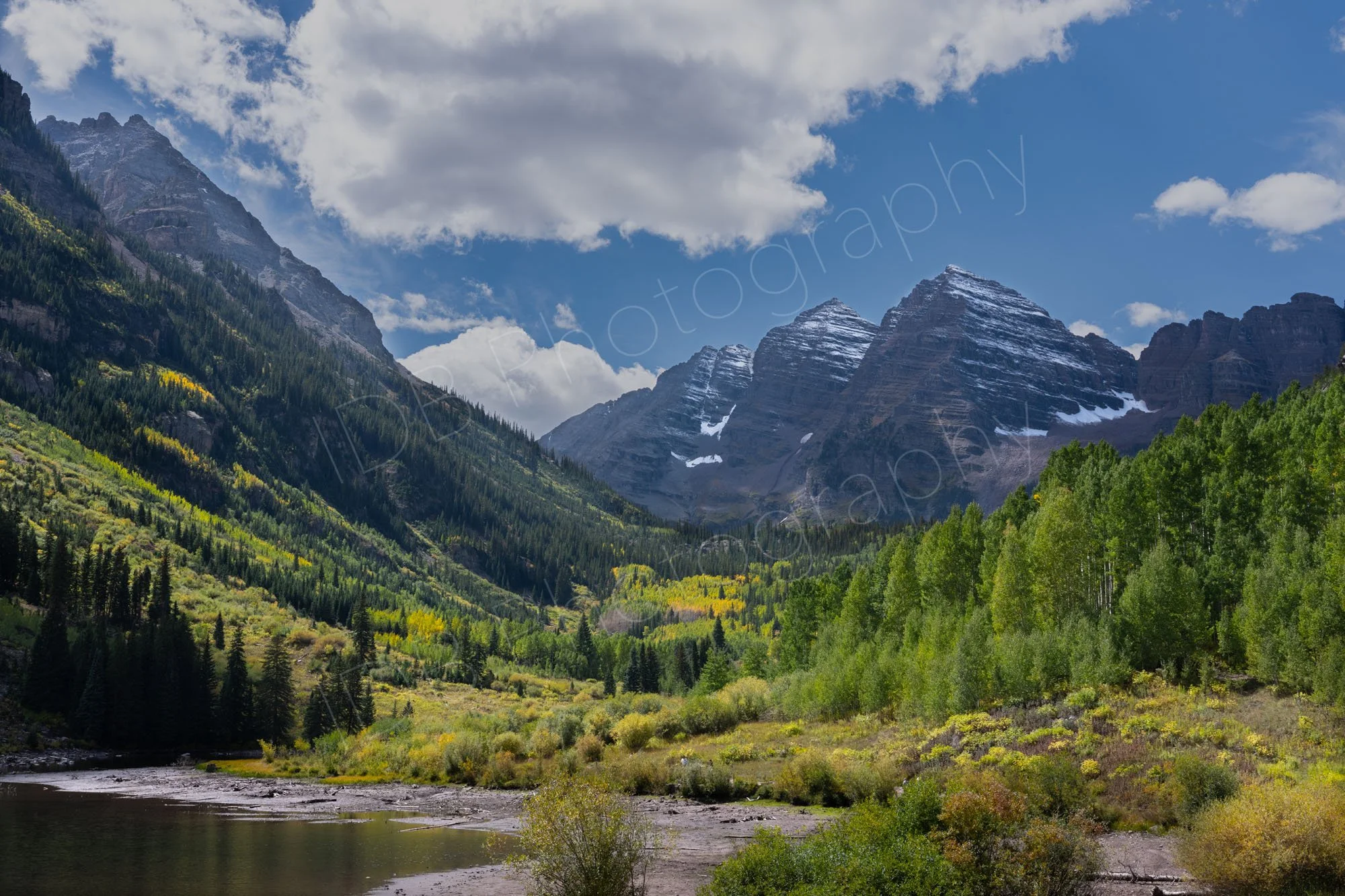 Maroon Bells Lake, Colorado and surrounding mountains with snow residue.  Conifers with green and yellow Aspen trees fill the valley floor; blue sky and clouds. 2.