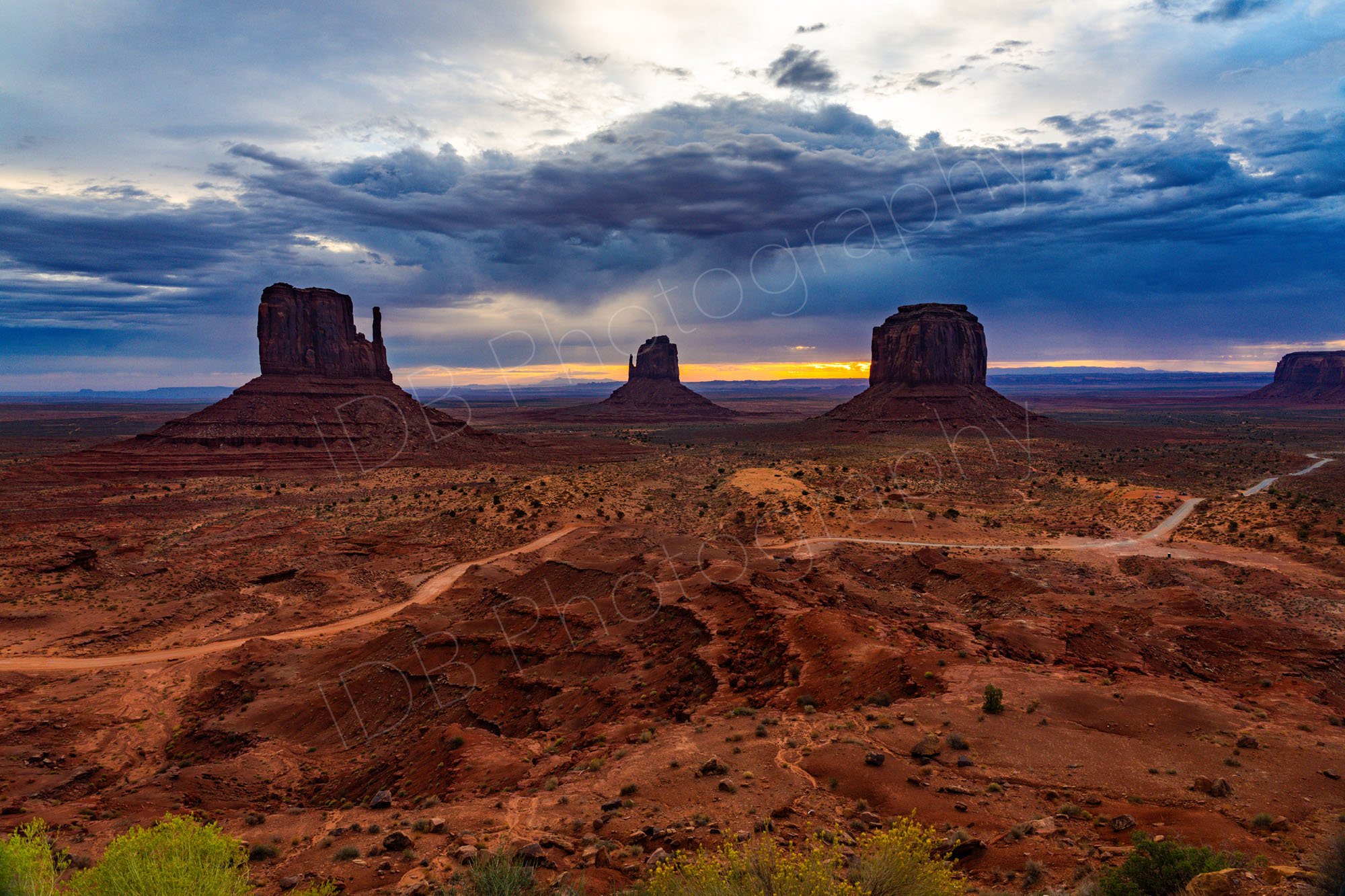 West Mitten, East Mitten and Merrick Buttes in Monument Valley with desert foreground and dramatic cloudy sunrise sky.