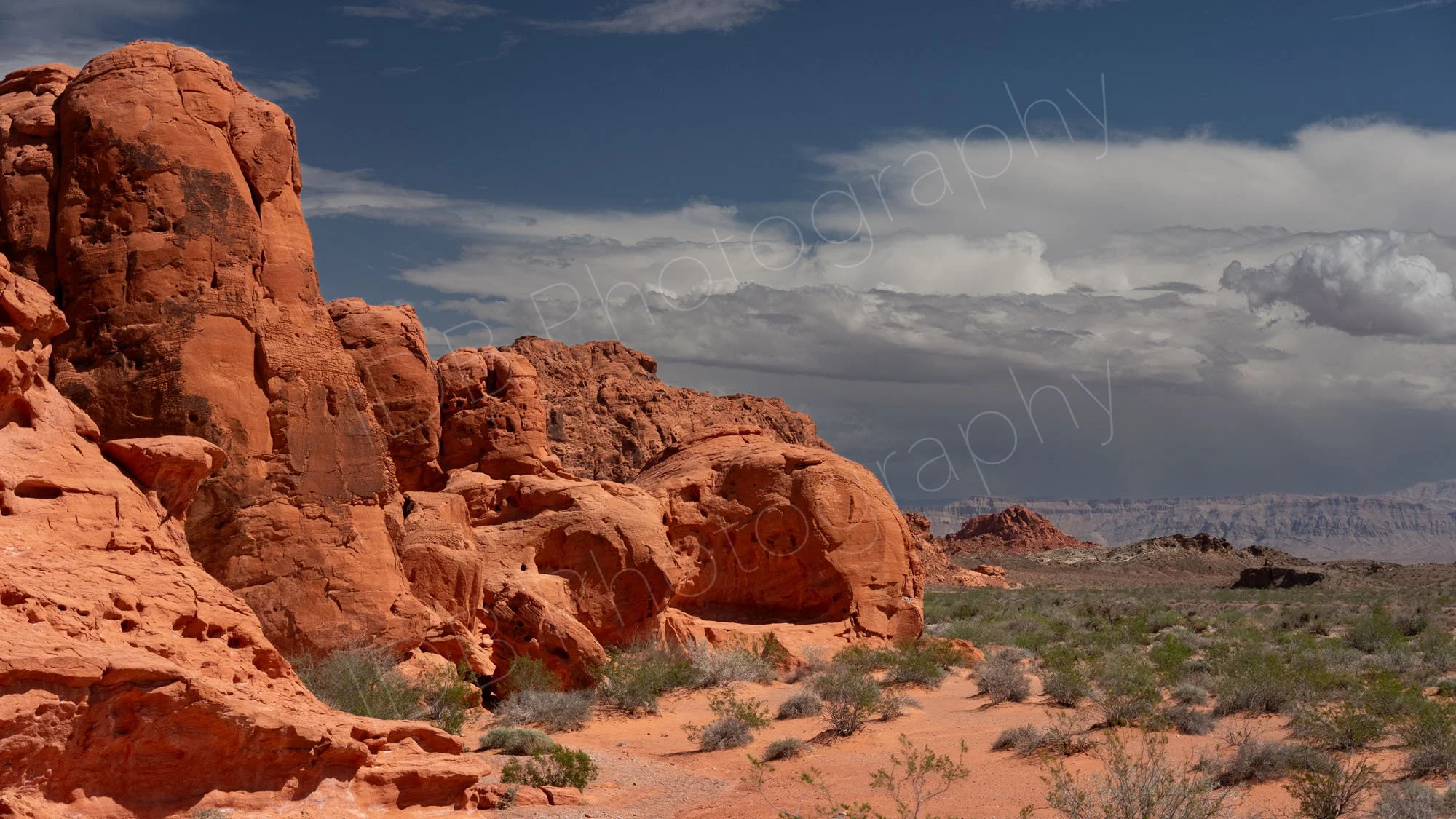 A large cluster of irregular red Aztec Sandstone with scrubby desert and cloudy blue sky background in Valley of Fire State Park, Nevada.