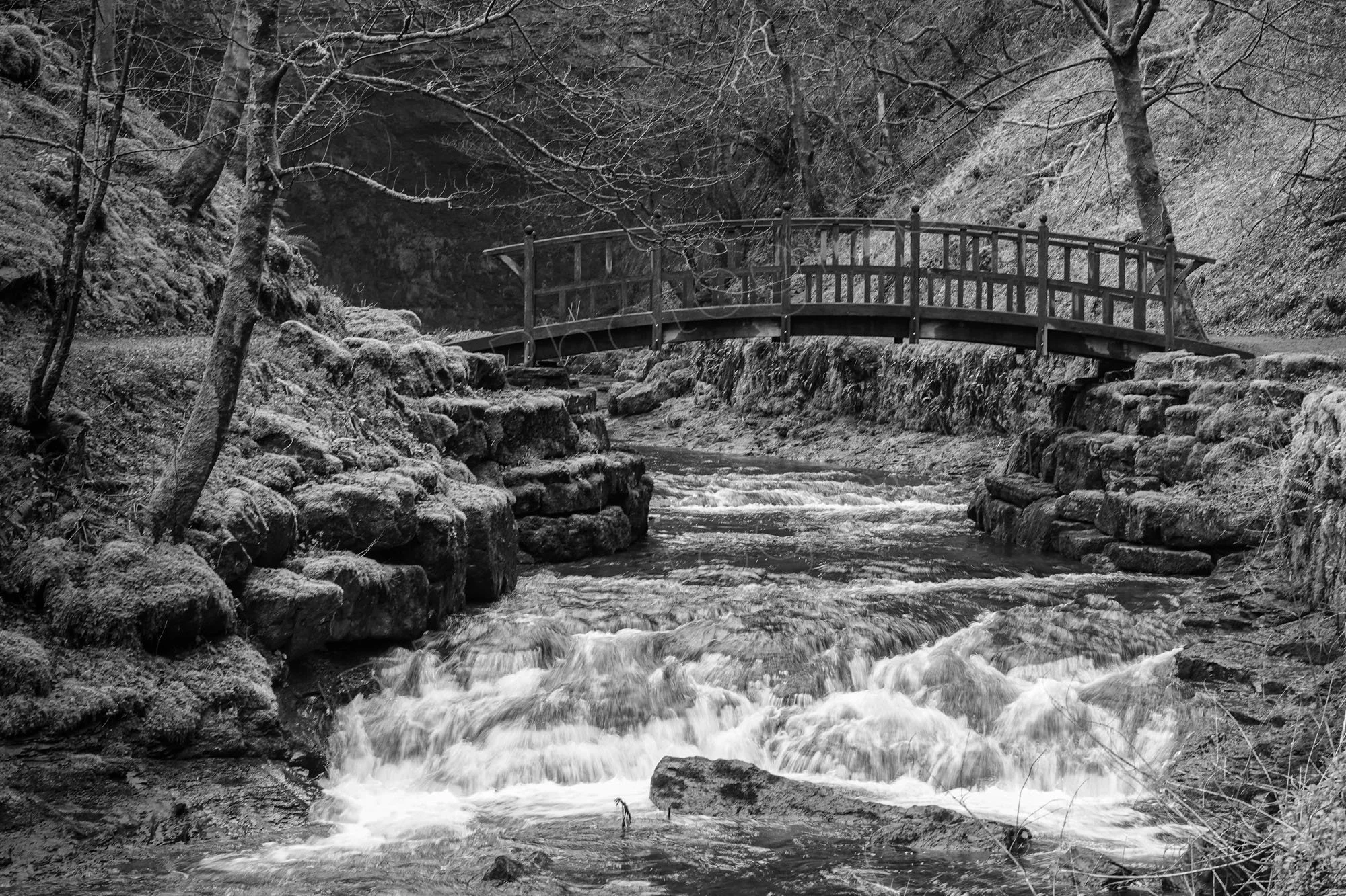 A black-and-white image with a rustic wooden arch bridge spanning a rushing stream with cascading small waterfalls over textured, mossy rocks.