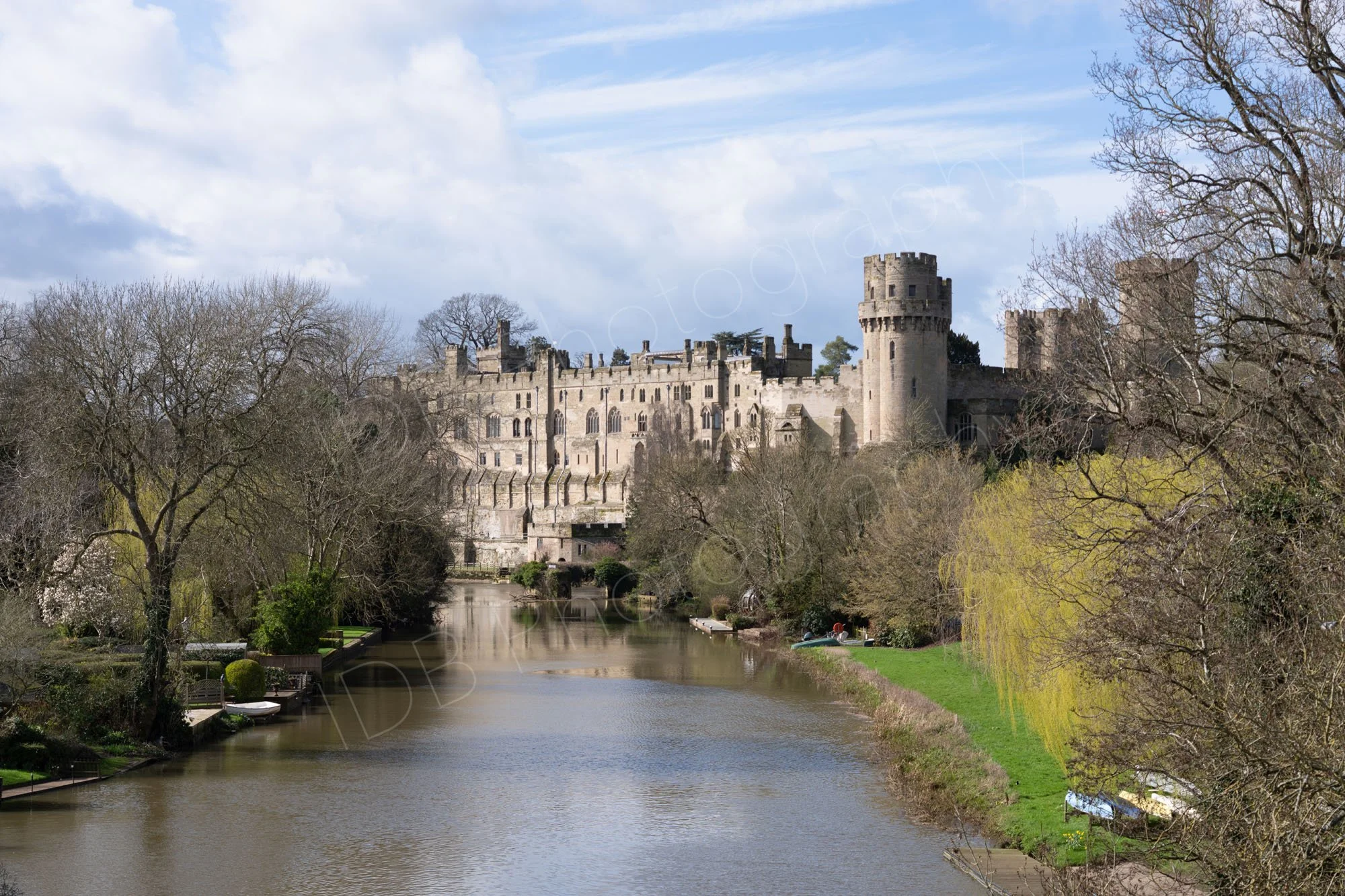 A view of Warwick Castle along the River Avon in early spring, some trees in leaf, others still bare, blue cloudy sky.