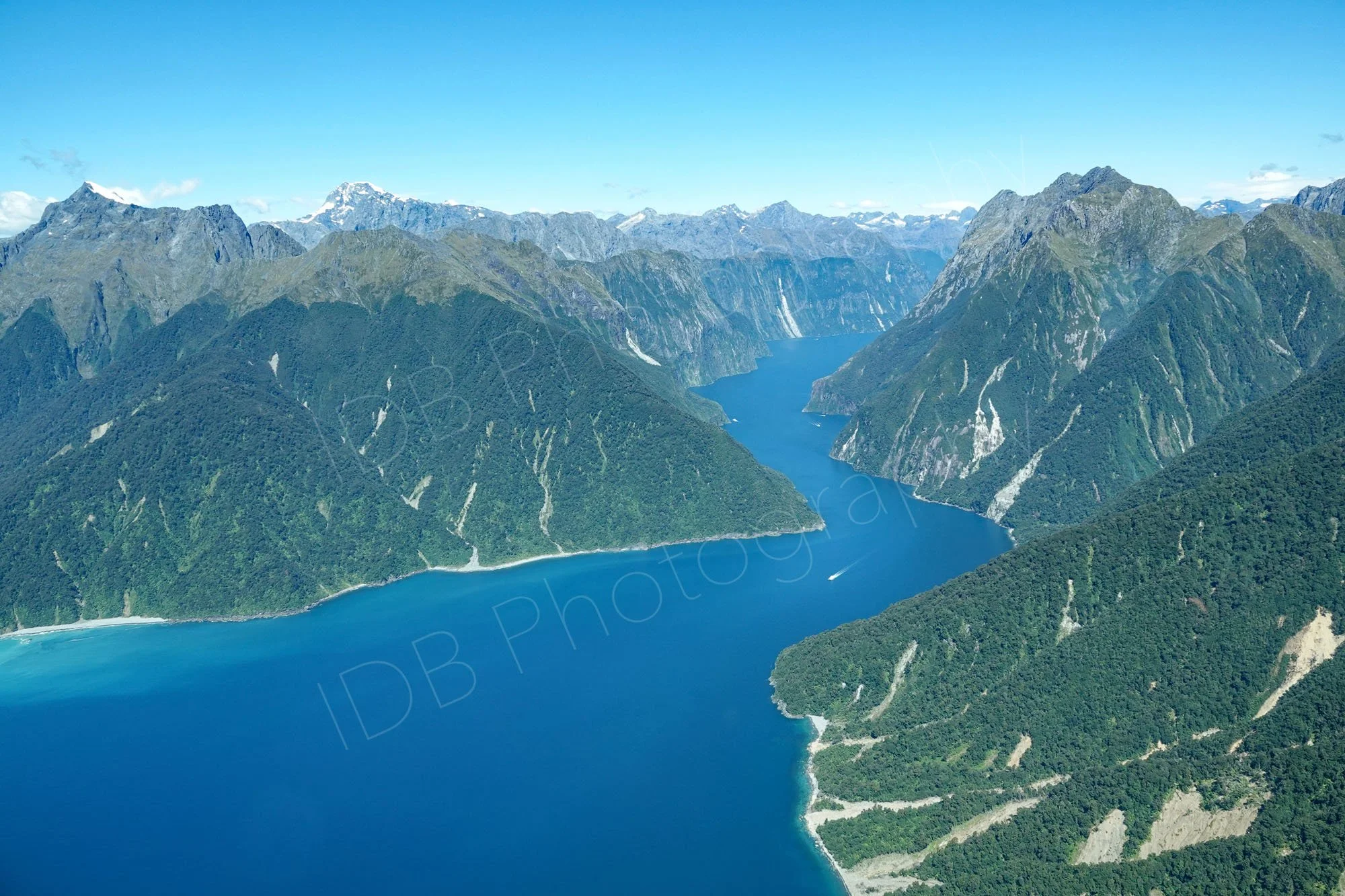 Aerial view of blue waters of Milford Sound, Fiordland, Southlands, NZ, steep vegetation covered mountains with snow capped peaks in the distance.