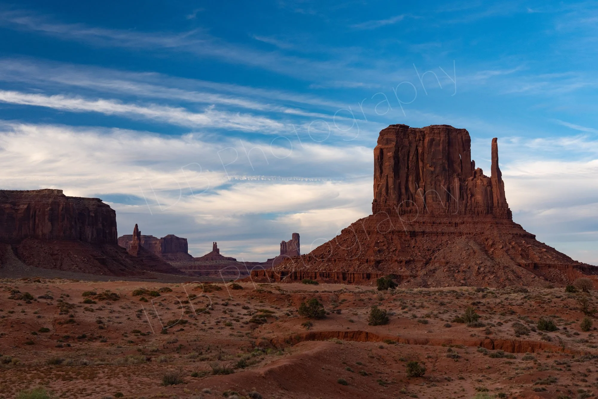 A scenic view of sandstone buttes and mesas in Monument Valley Navajo Tribal Park, Utah, under a cloudy blue sky, West Mitten butte