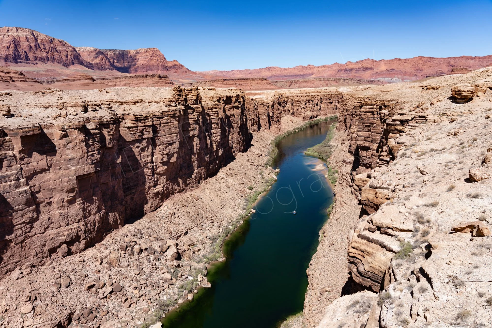 A view from the Navajo Bridge of the layered cliffs and Colorado River at Marble Canyon, Arizona; the surrounding red mountain landscape is seen with blue sky.