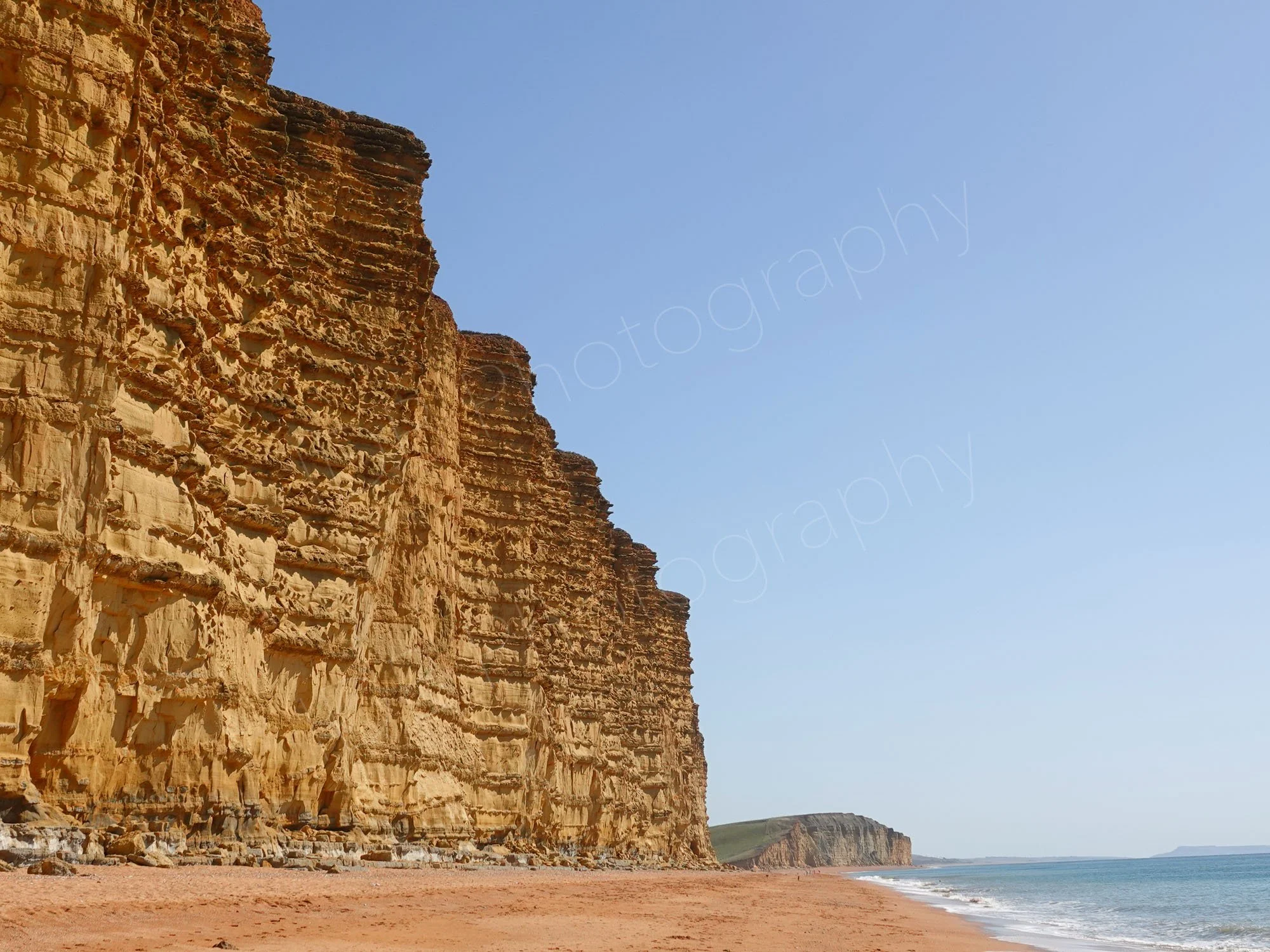 The striking, vertical golden-yellow banded sandstone cliffs at West Bay in Dorset, with calm sea under a blue sky.