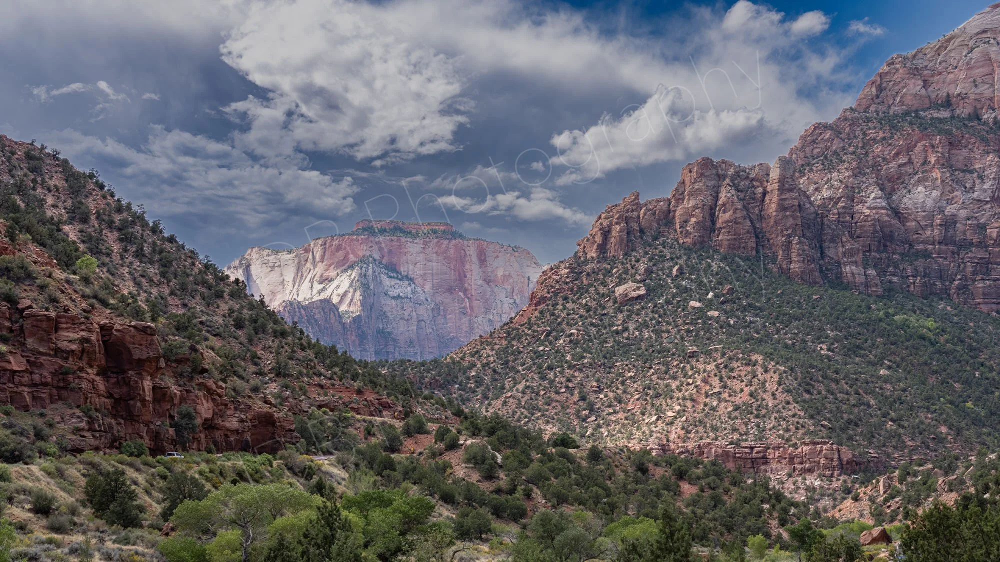 Red rock cliffs, green valley brush, and the prominent mass of The East Temple in the background under a cloudy sky at Zion National Park.2.