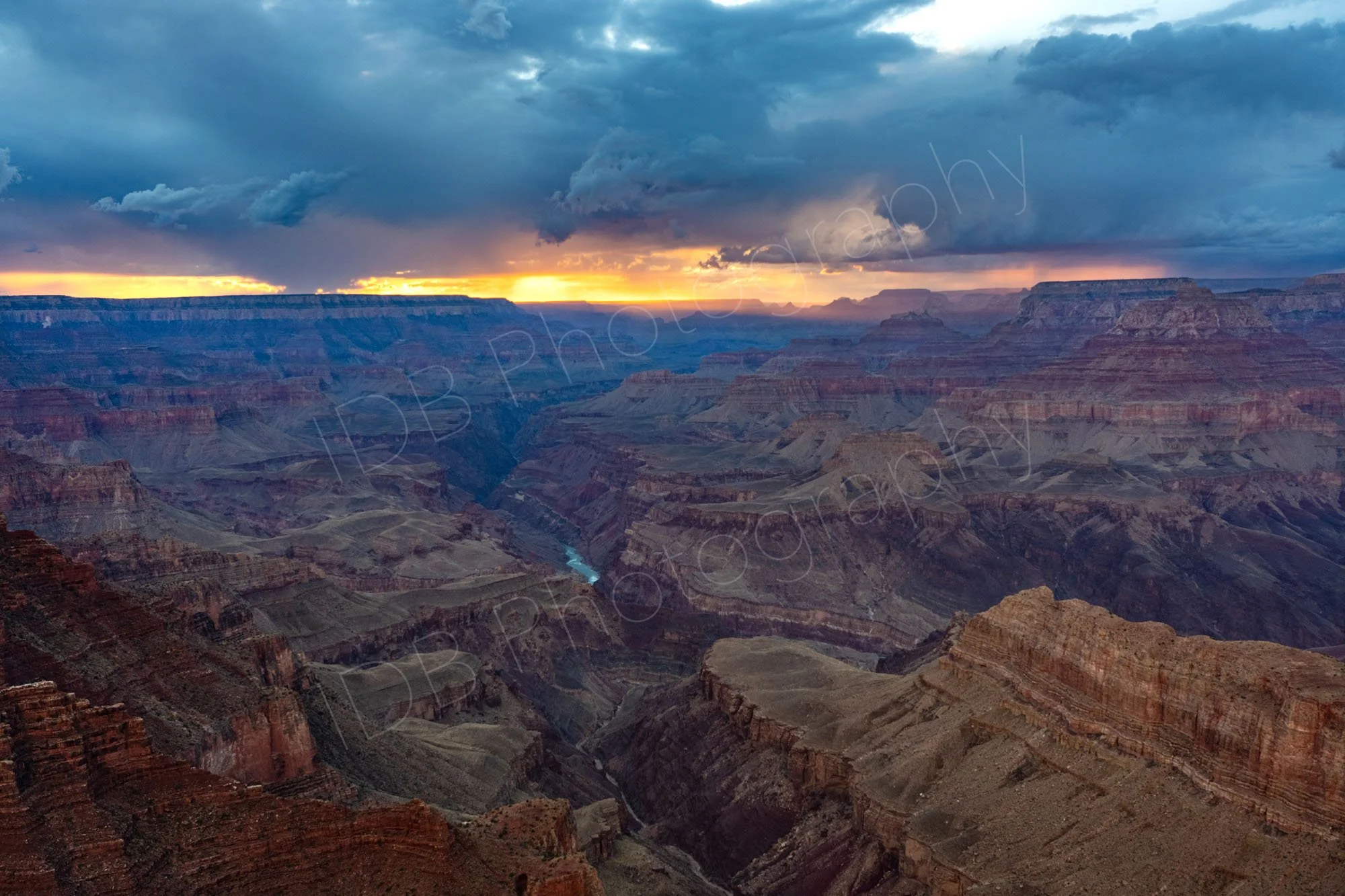 A wide expanse sunset view over cliffs and gorges of the Grand Canyon, Arizona around the Colorado River, with storm clouds above.