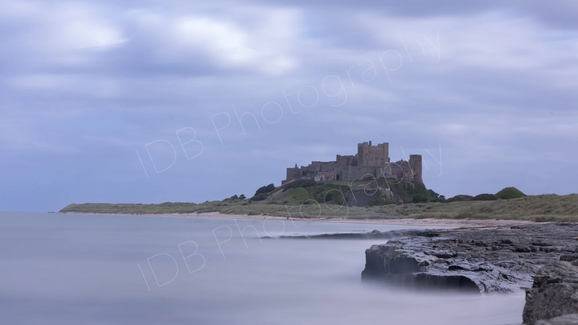 A long exposure image of Bamburgh Castle, Northumberland and rocky shoreline, with silky smooth sea, and blue/grey cloudy sky.