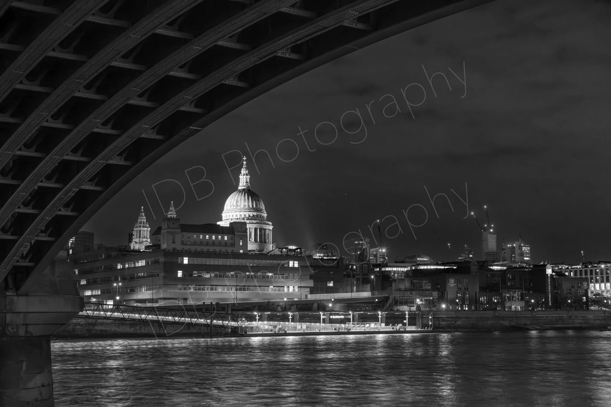 A black and white late evening view of St. Paul's Cathedral framed underneath the structure of Blackfriars Railway Bridge across the River Thames in London.