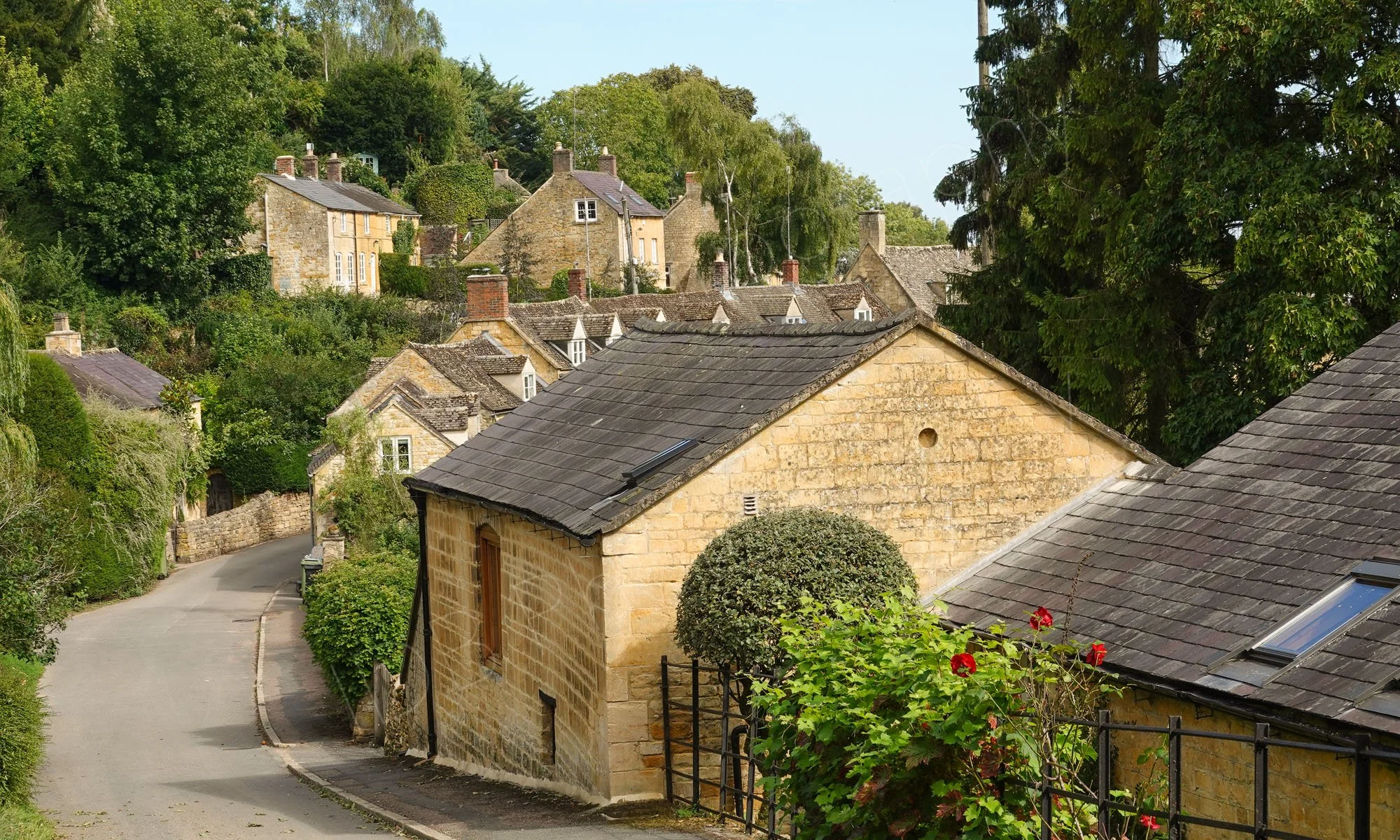 A view of part of the High Street in Blockley, a beatiful village in the Cotswolds. A narrow road with Cotswold stone buildings and blue sky.