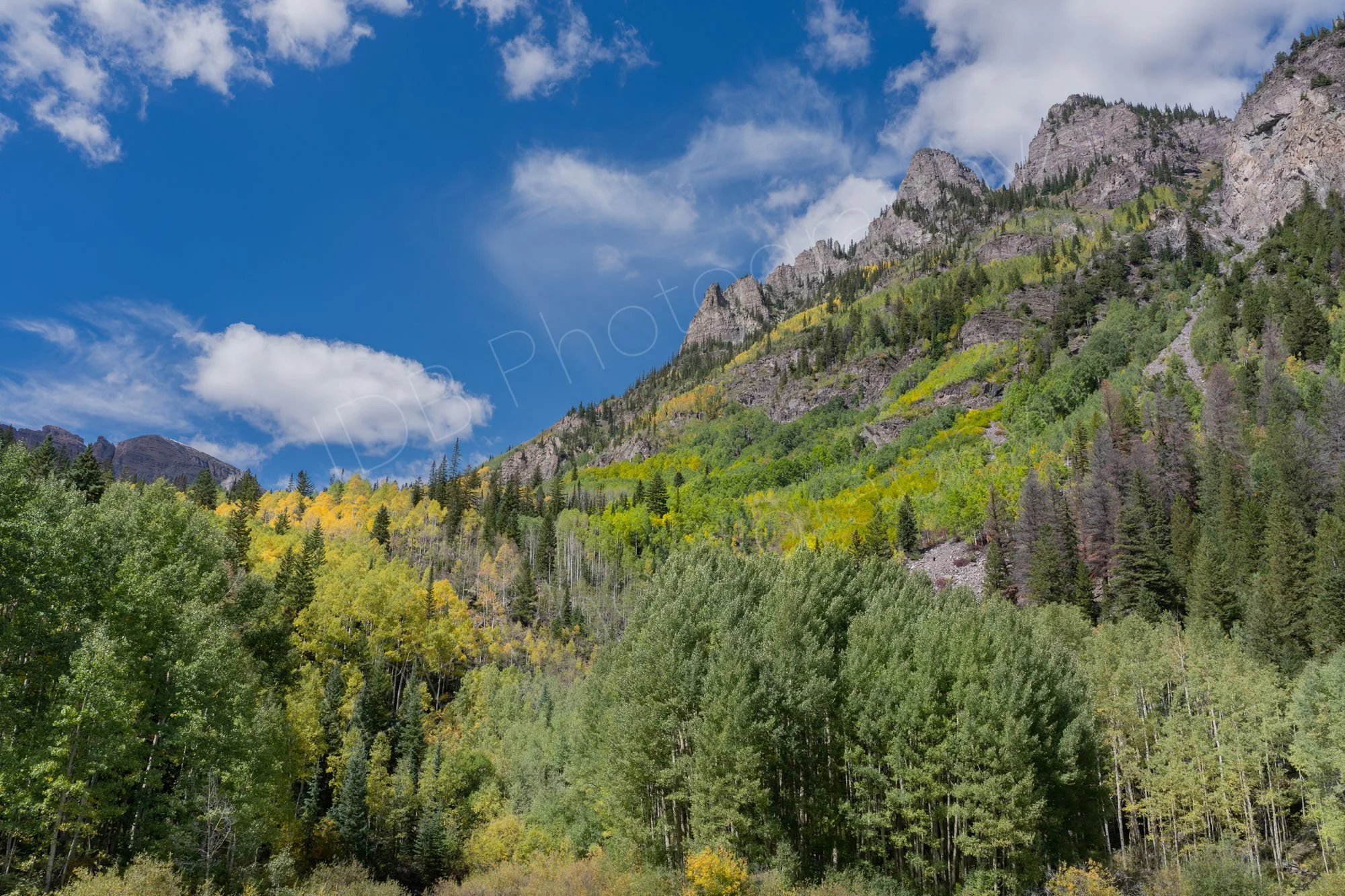 A view of the mountains, pines and green and gold aspen trees at Maroon Bells Lake with a cloudy blue sky.
