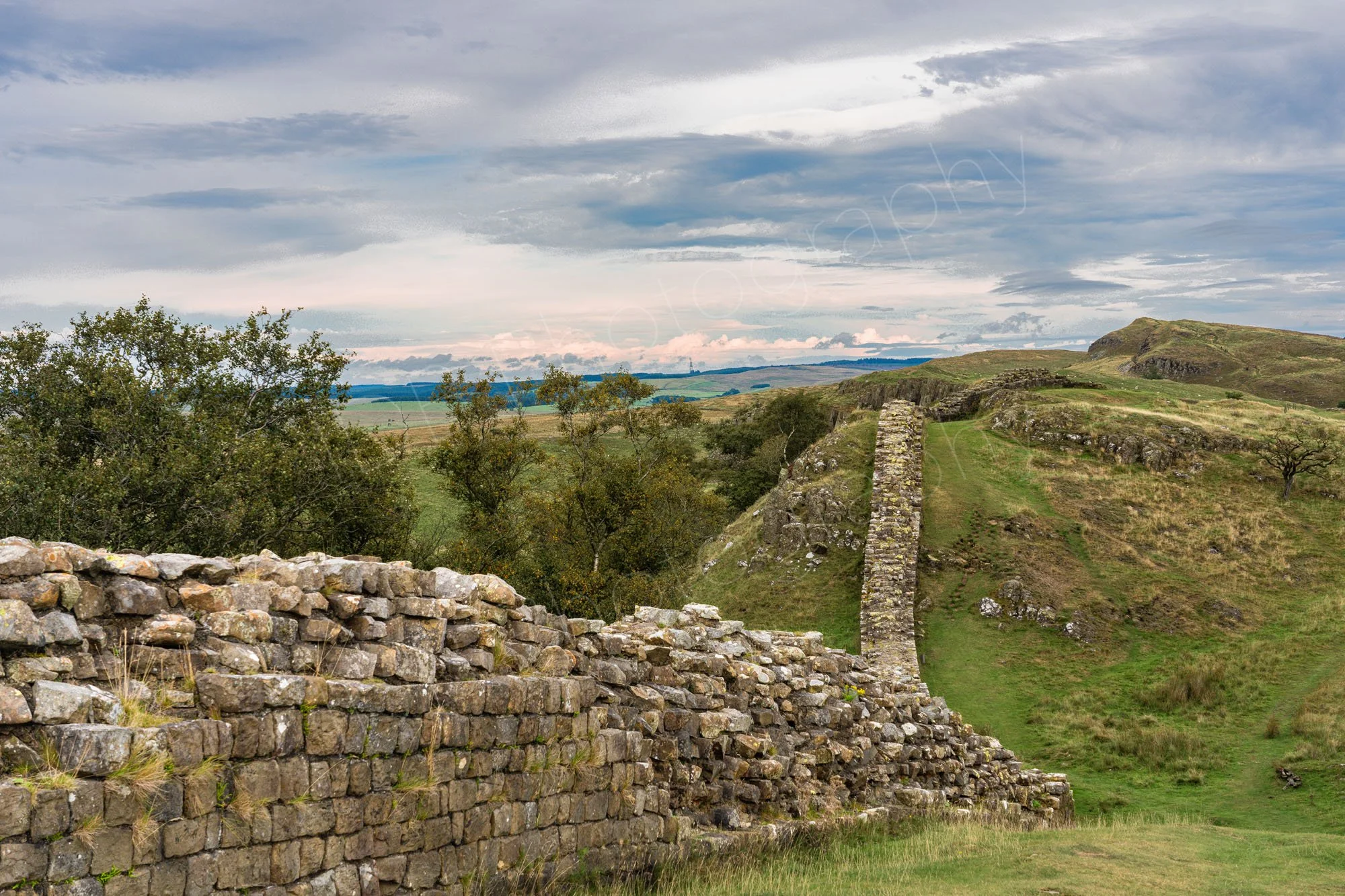 Hadrian's Wall near Walltown crags, Greenhead, Northumberland, hilly terrain, trees, greyish blue cloudy sky.