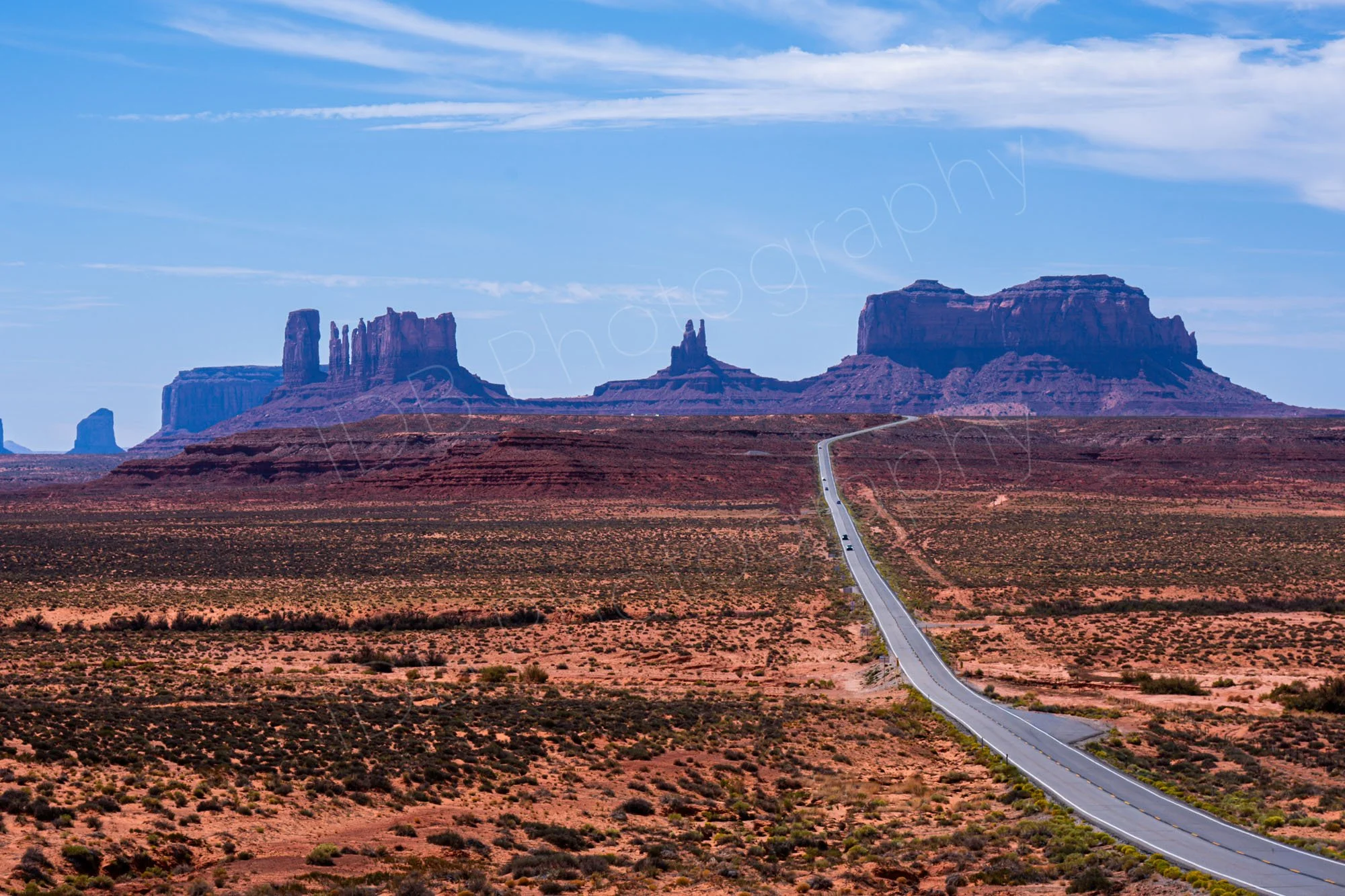 A view towards Monument Valley from near Forest Gump point on US Route 163 which leads through the desert to the massive buttes, cloudy blue sky background.