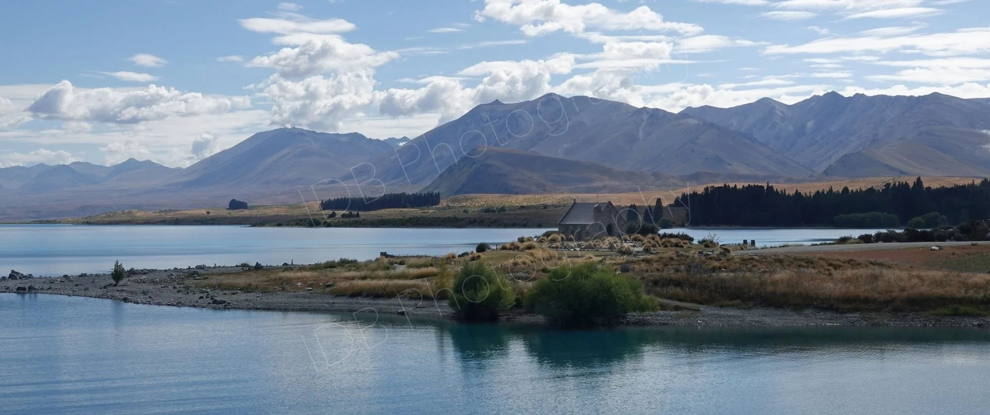 Panoramic type view of Lake Tekapo and the Church of the Good Shepherd, mountains and bright cloudy sky in the background. Canterbury, New Zealand.