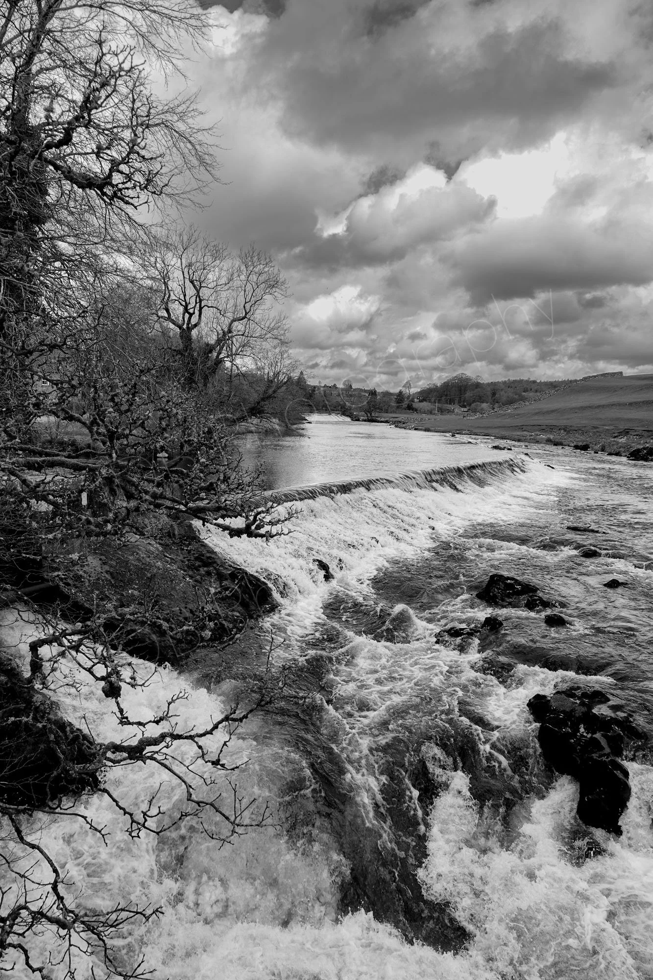 A black-and-white image of the cascading weir and rushing, foamy water over the limestone rocks at Linton Falls, North Yorkshire, dramatic cloudy sky.
