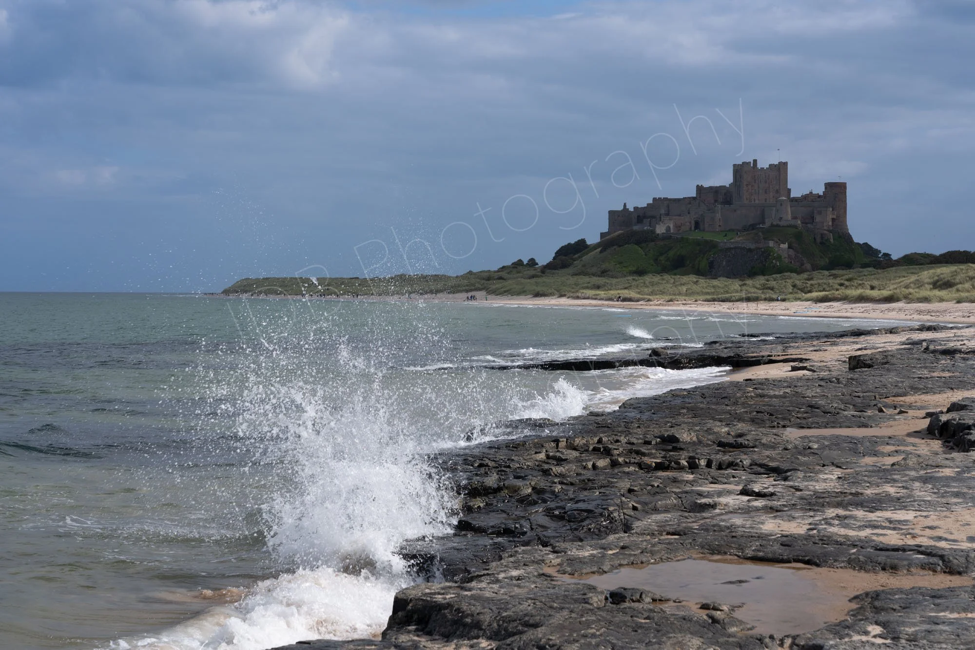 Bamburgh Castle, Northumberland and rocky shoreline, with breaking waves splashing on the rocks, grey blue cloudy sky.