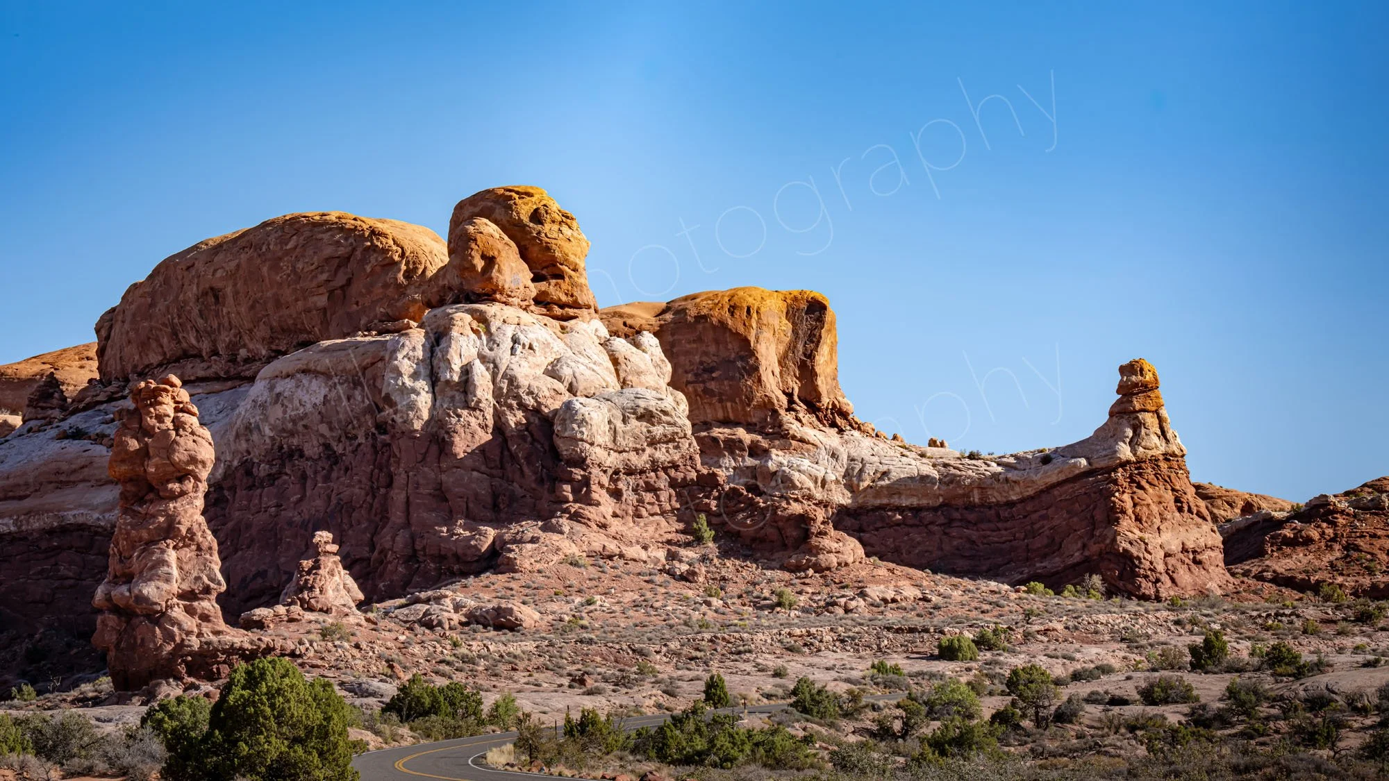 The Sphinx and Rhinoceros rock formation at Arches National Park, Moab, Utah with a road and scrubby desert foreground and blue sky.