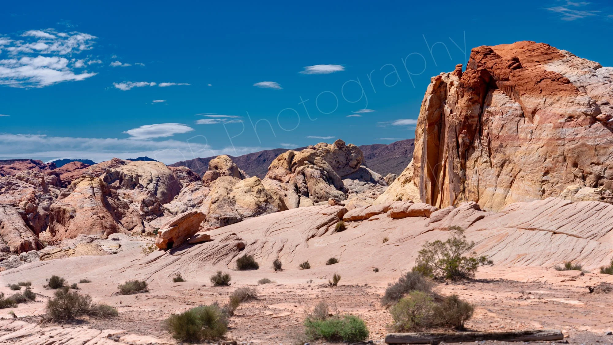 An irregular landscape of various buff coloured and red rocks with a scrubby desert foreground and cloudy blue sky background in Valley of Fire SP, Nevada