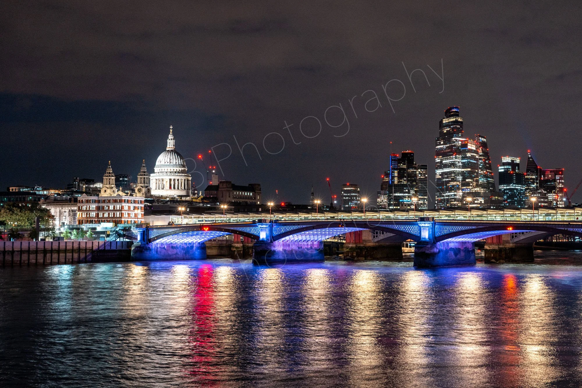 An illuminated night view of the River Thames  featuring Blackfriars Railway Bridge, with St Paul's Cathedral and the City of London skyline; nice reflections on the water.