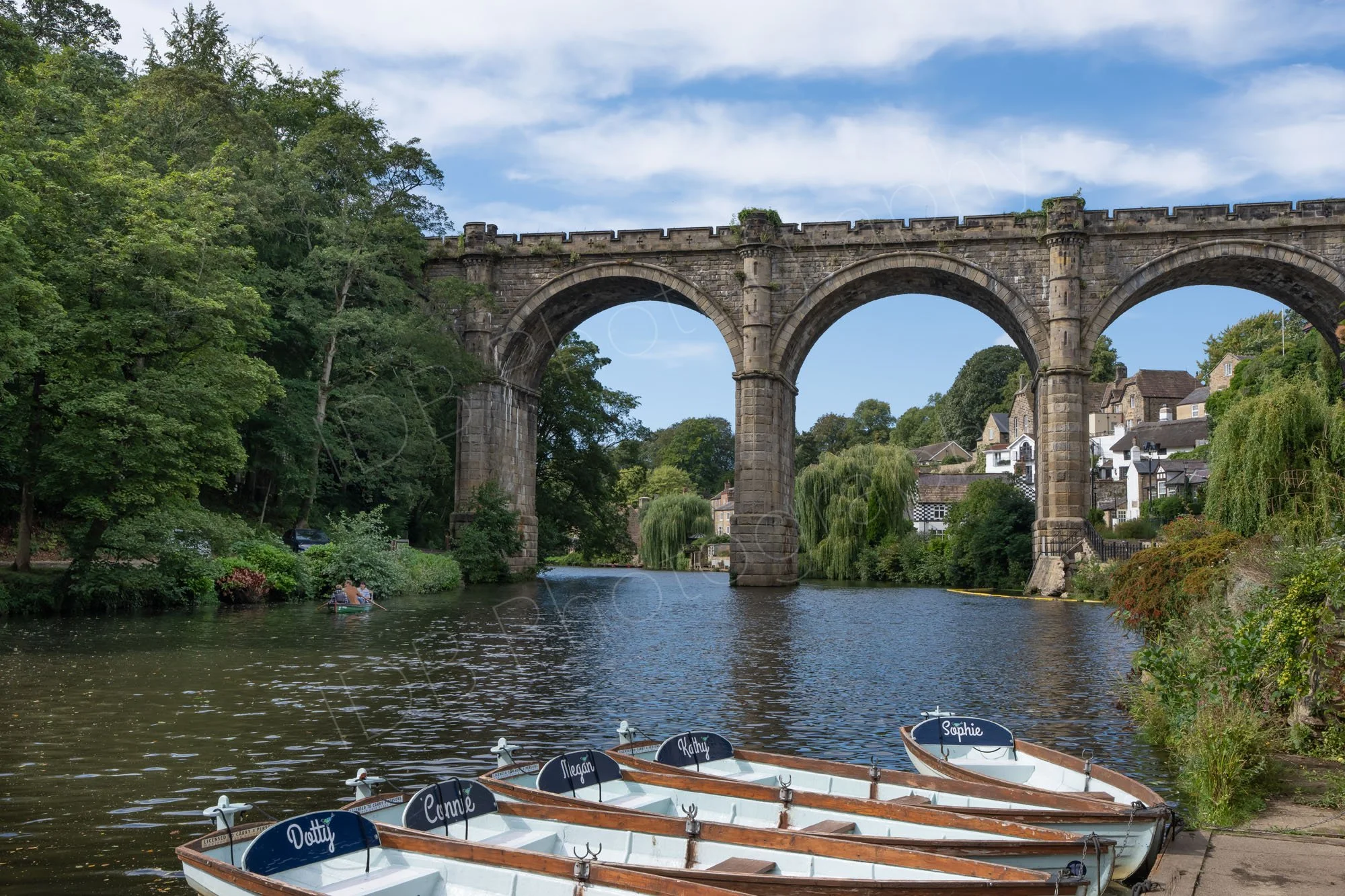 View towards the Knaresborough, Yorkshire viaduct over the River Nidd with named hire boats in the foreground, green trees, cloudy blue sky.