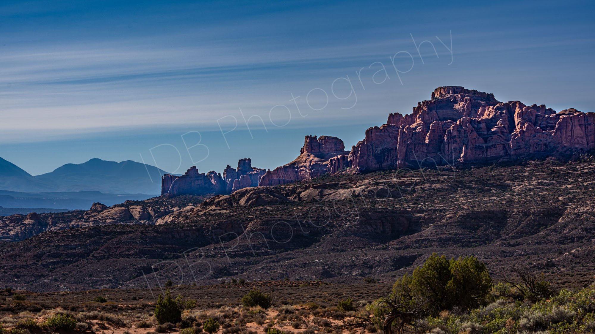 A mass of rugged red rocks rise from the desert in Arches National Park with an interesting streaky cloud blue sky.