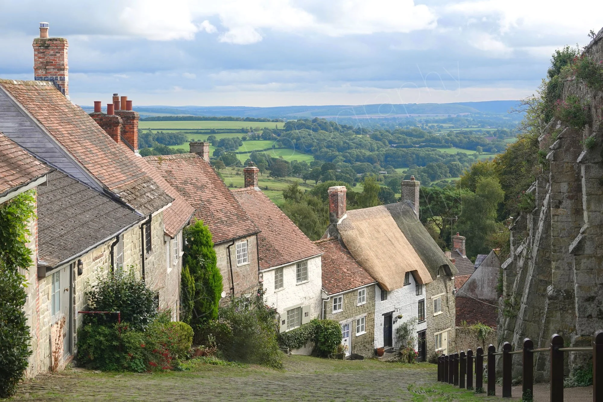 The steep cobbled Gold Hill lined by old stone cottages and thatched houses in Shaftesbury, Dorset, England, with countryside and cloudy sky behind..