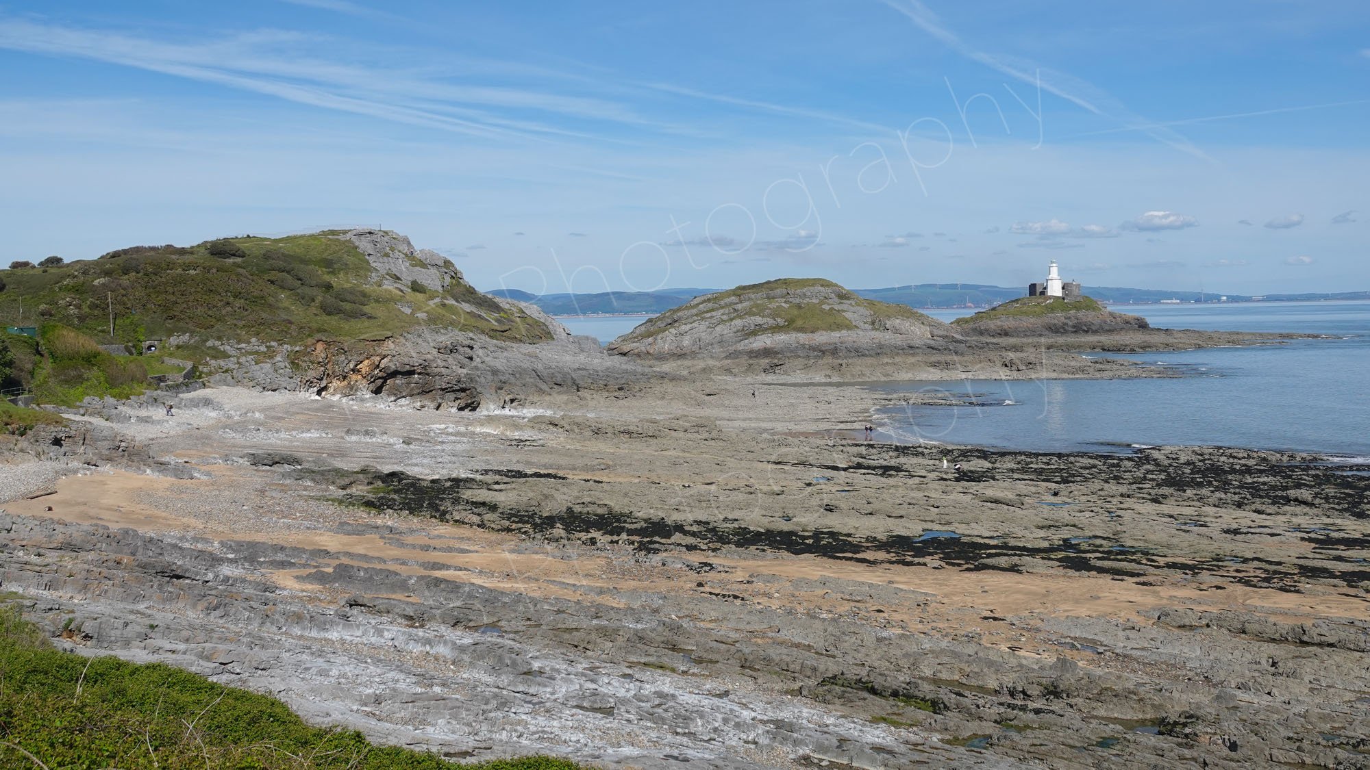A view of the rocky Bracelet Bay and the Mumbles Lighthouse on the  edge of the Gower Peninsula, Swansea; cloudy blue sky.