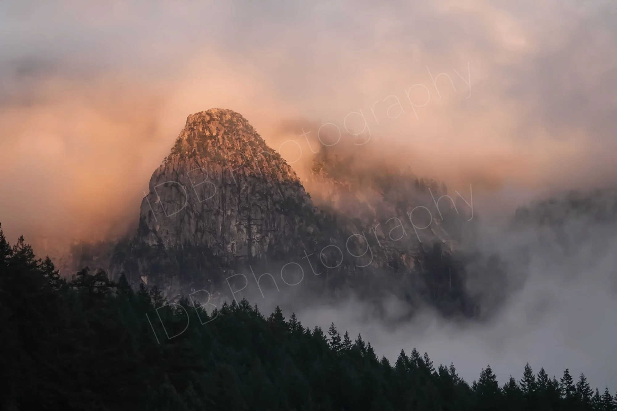 A rocky peak (? Slhanay Peak)  rising out of the dense forest amid an orange sunset mist, near Squamish, British Columbia.