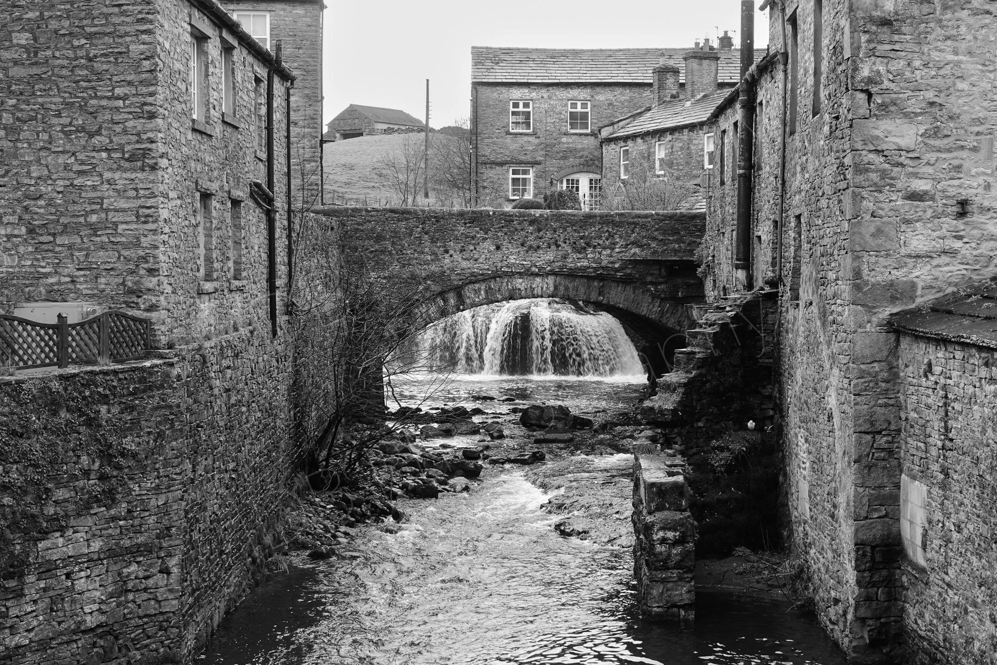 A black and white image of a waterfall  framed by a stone bridge arch with surrounding stone buildings in Hawes, N. Yorkshire.