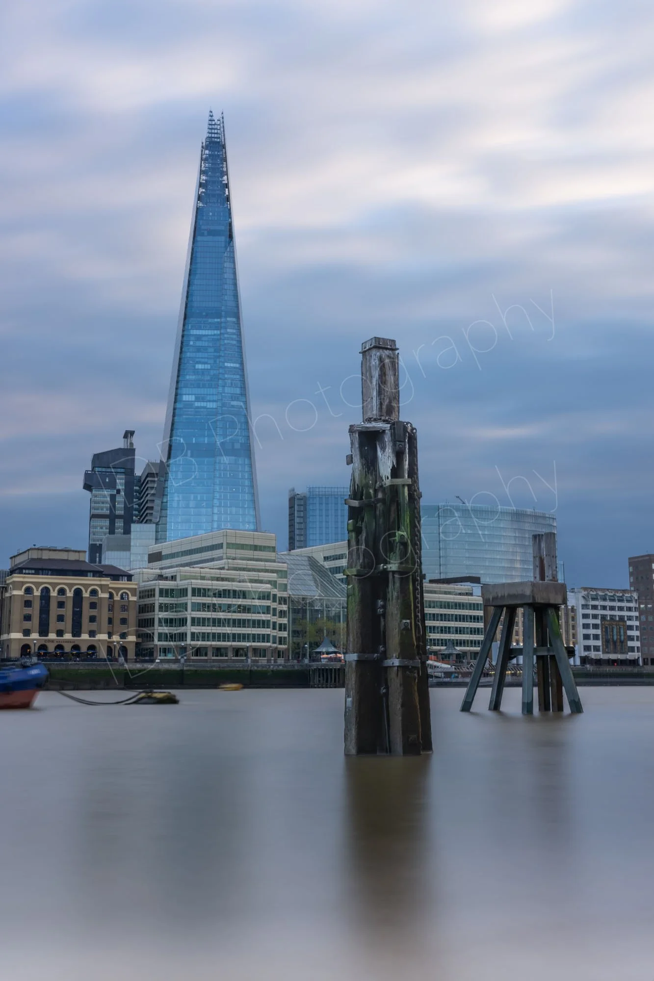 A long exposure ND filter image across the Thames. Old wooden jetty posts protrude from the still water with the Shard behind beneath an eerie cloudy blue sky.