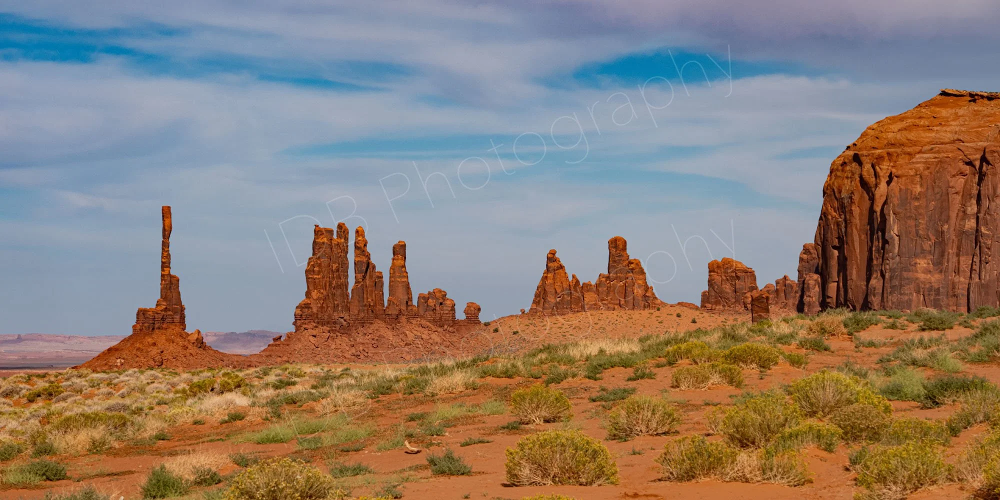 The famous Totem Pole and the adjacent Yei Bi Chei rock spires in Monument Valley Navajo Tribal Park, sparse desert foreground with cloudy blue sky.