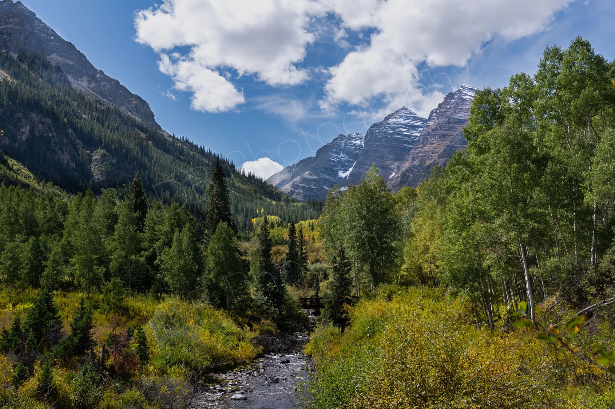 A stream at the base Maroon Creek Valley, surrounded by shrubs, pine and aspen trees with mountains dusted with snow, cloudy blue sky.