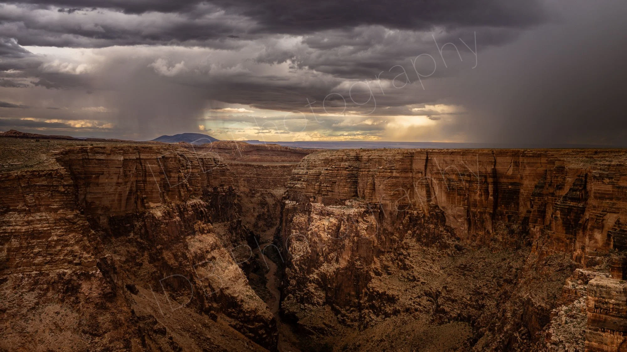 Deep, rocky canyon of the Little Colorado River Gorge, Arizona, USA, with layered sandstone cliffs beneath a dramatic, stormy sky with rain shafts.