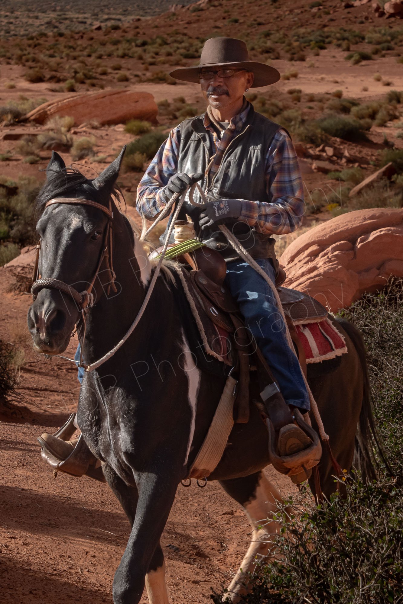 A male Navajo wearing wide-brimmed hat, a plaid long-sleeve shirt, and a dark gilet, rides his horse in Monument Valley Tribal Park, Utah.