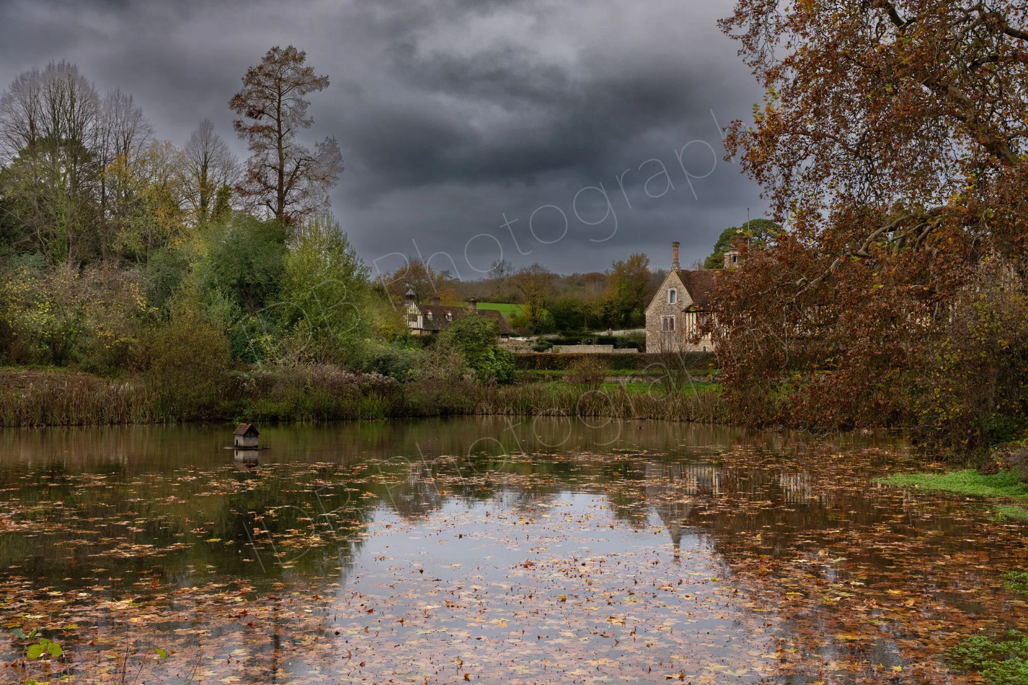 A view across the lake at Ightham Mote, near Sevenoaks, Kent, on an autumn day with both reflections and leaves on the water, storm clouds behind.