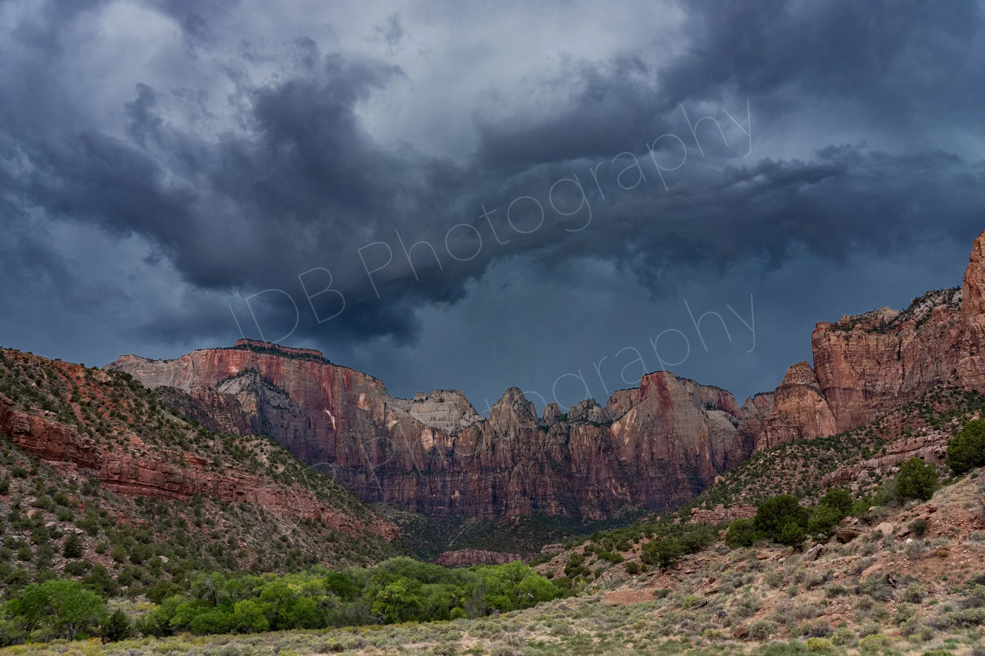 An "After the Storm" image of red and brown cliffs of Zion Nationa Park, Utah with scrubby desert and dramatic grey cloud patterns.