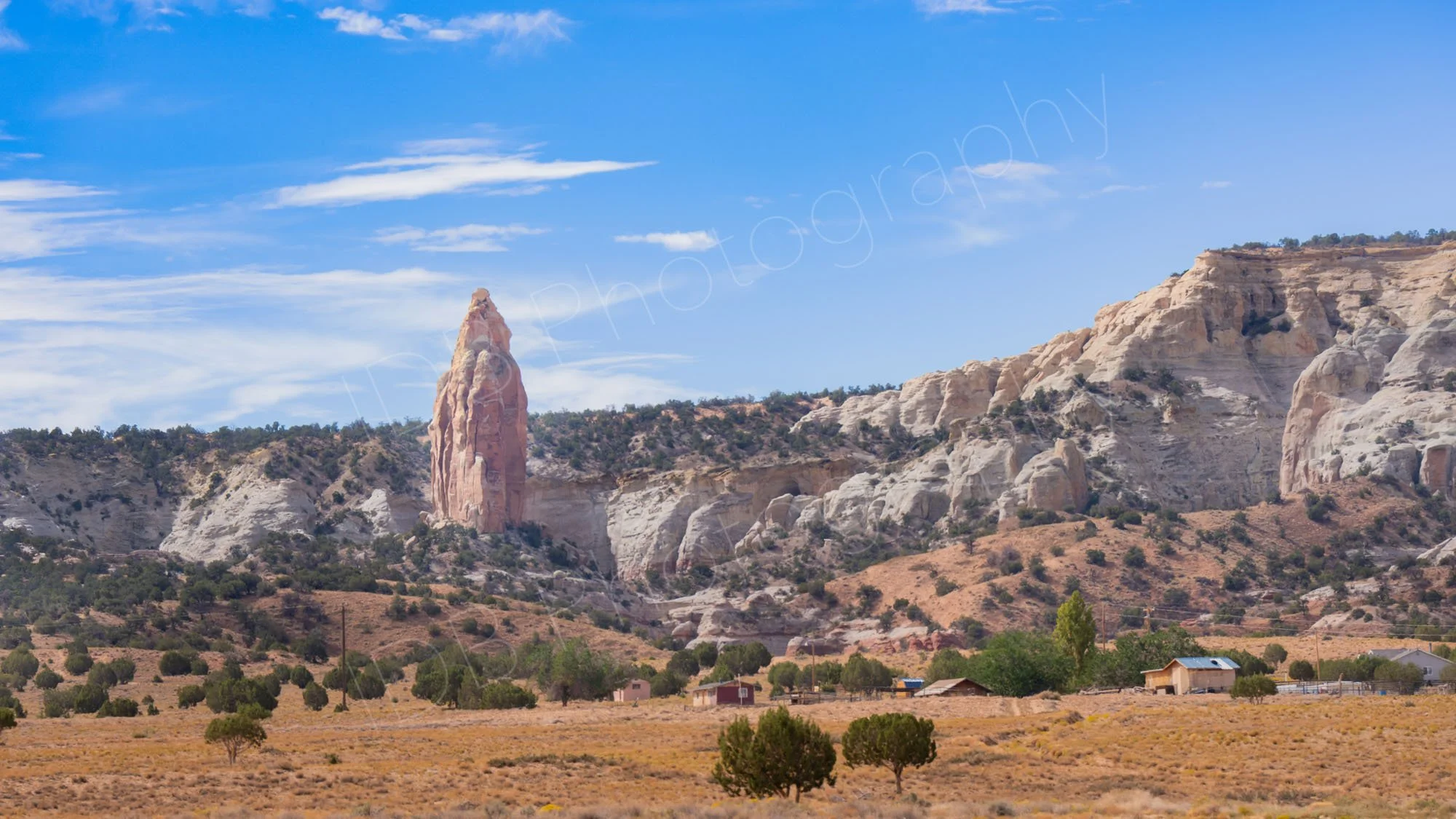 The pink spire of Chimney Rock, with white, buff cliffs Kodachrome Basin State Park, Utah, grassy foreground with trees, scattered buildings and blue sky.