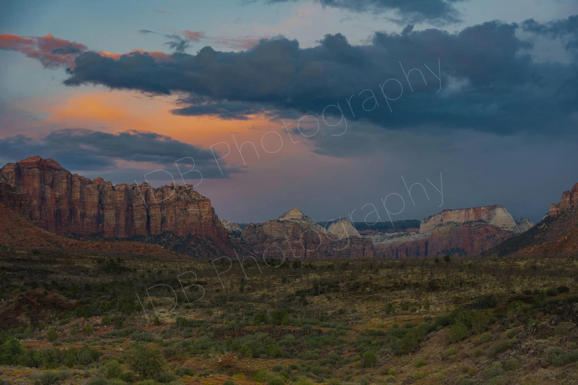 Mount Kinesava and other peaks with a green shrubby foreground, a beautiful orange sunset glow with storm clouds, Zion National Park, Utah.