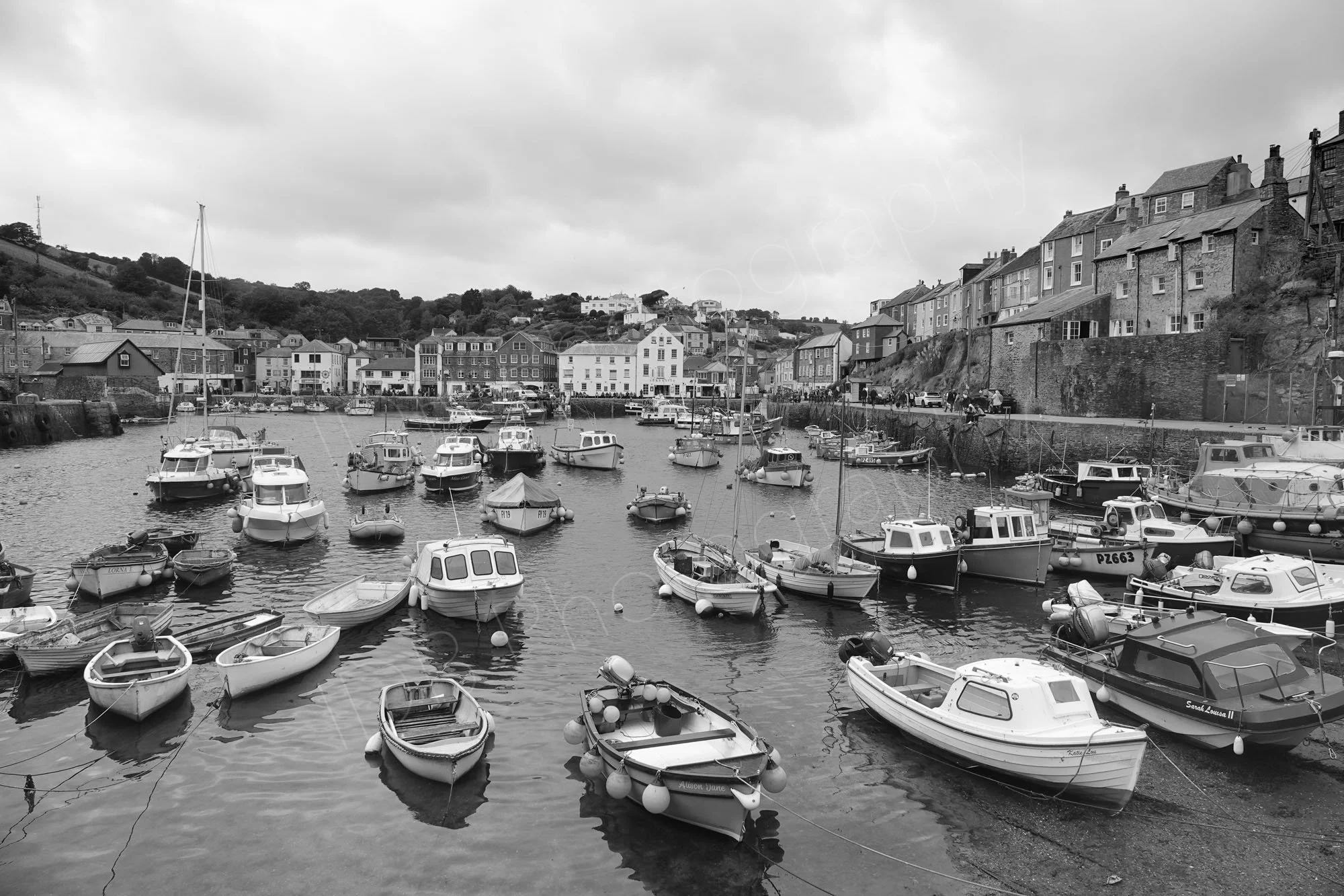 A black and white image of Mevagissy Harbour, Cornwall, with boats, houses and cloudy skies.