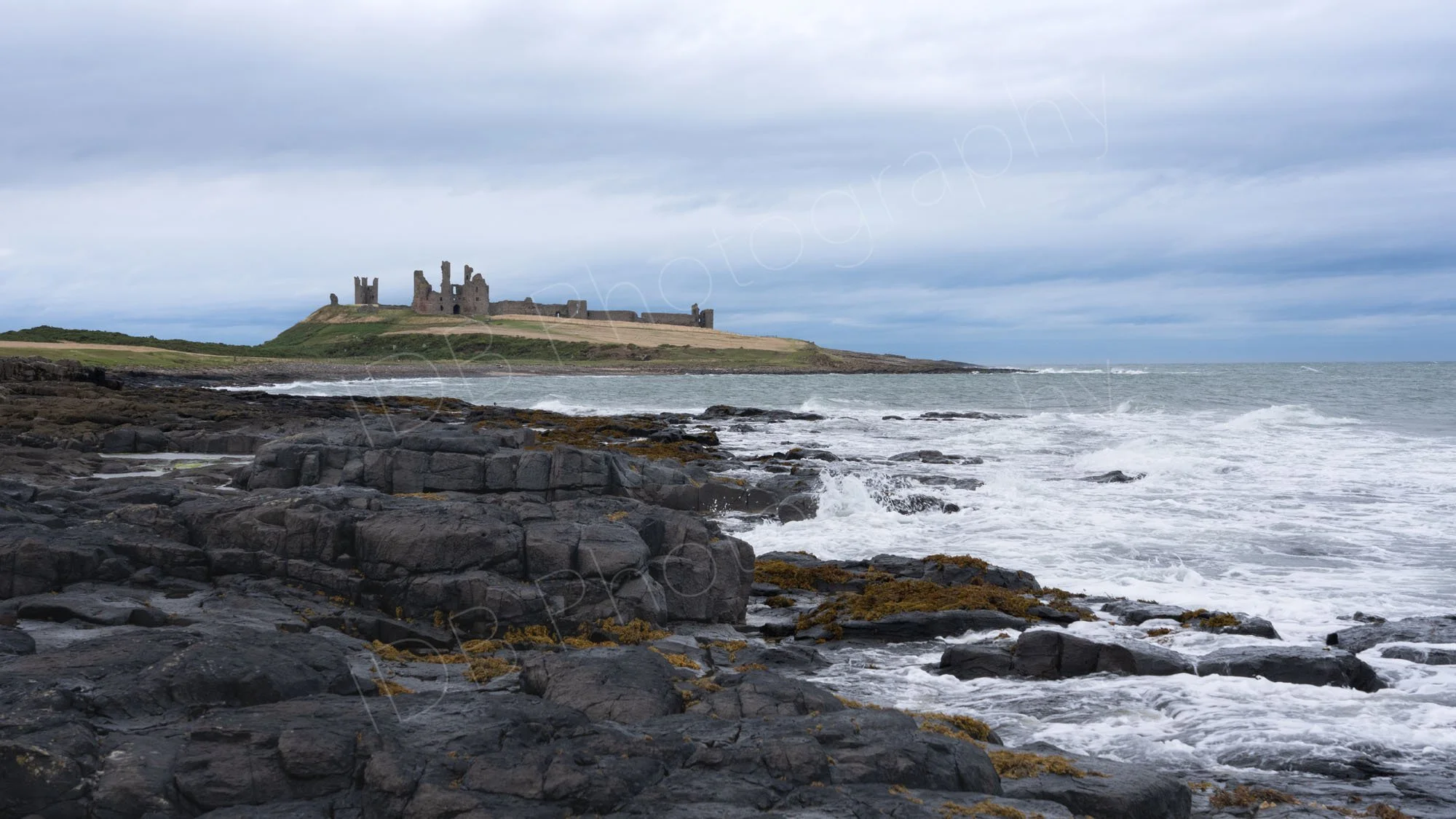 The ruins of Dunstanburgh Castle, Northumberland with a rocky foreground and breaking white waves, varied grey sky.