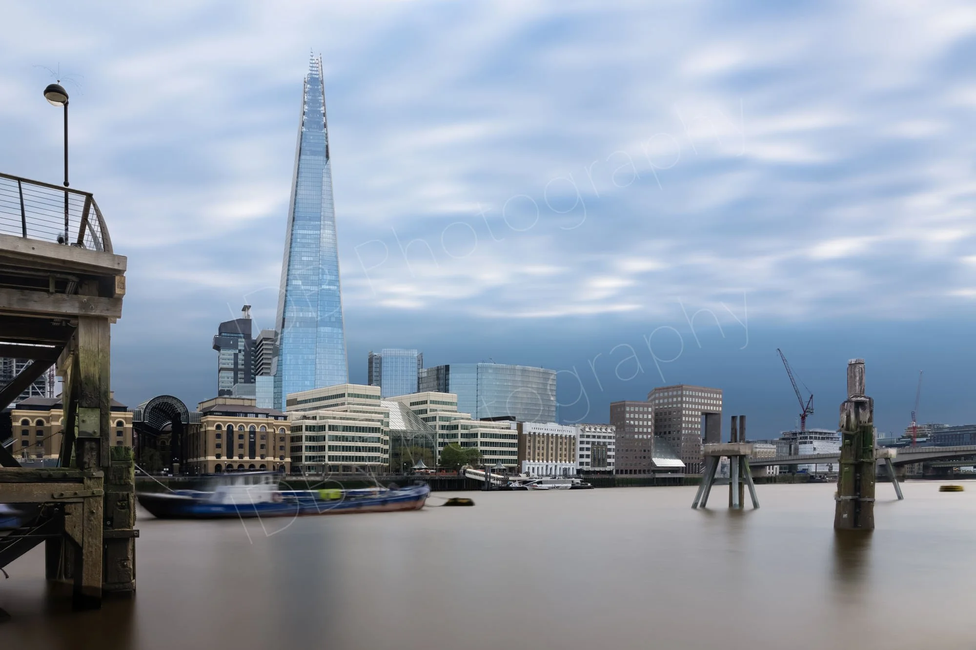 A long exposure ND filter image across the Thames. Old wooden posts project from the still water, a boat bobs eerily in front of The Shard, blue and white sky.