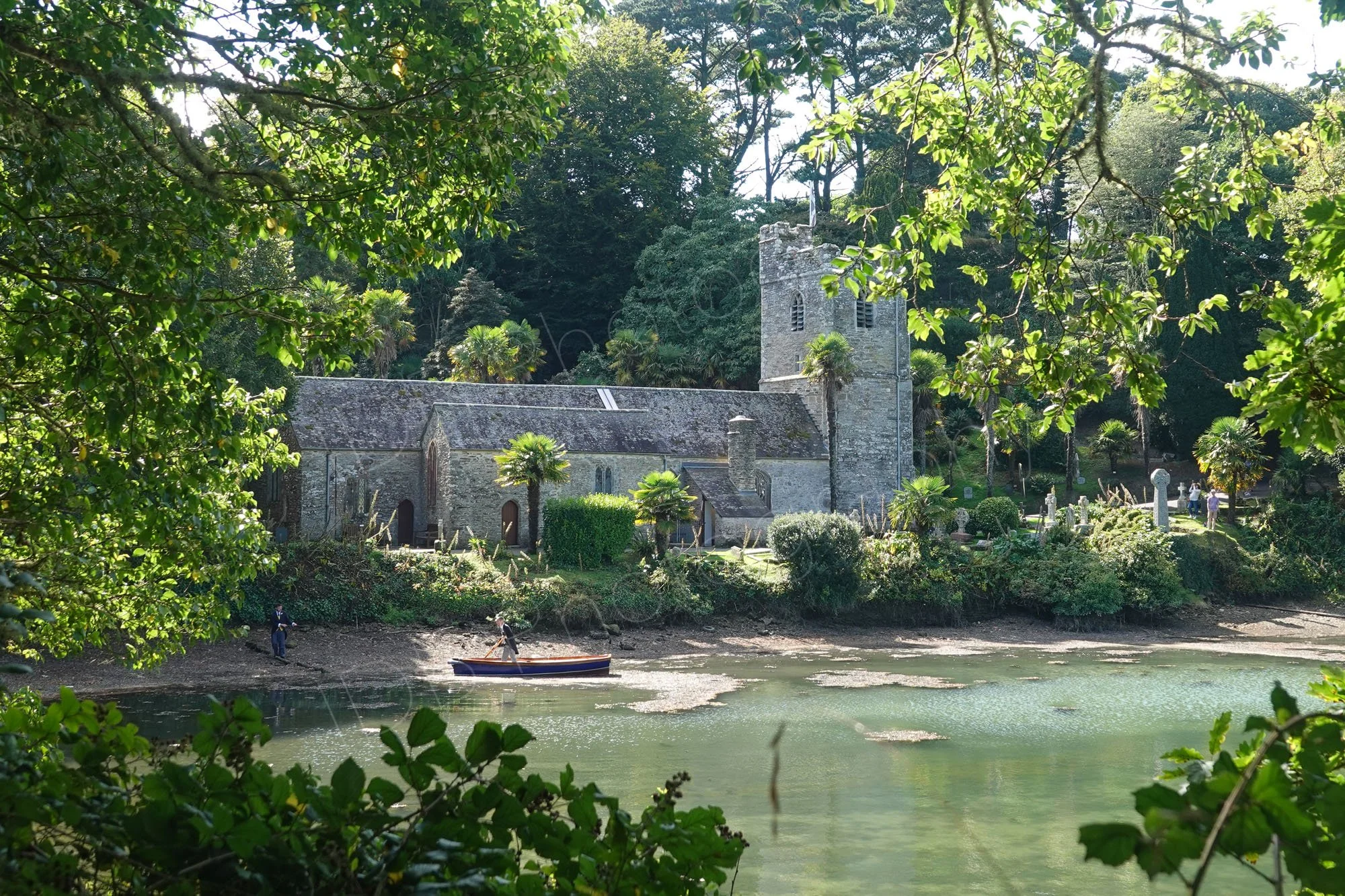 The historic St Just-in-Roseland Church situated beside the water in Cornwall, England. The image appears to show a Bridegroom and Best Man, arriving at the wedding by rowing boat.