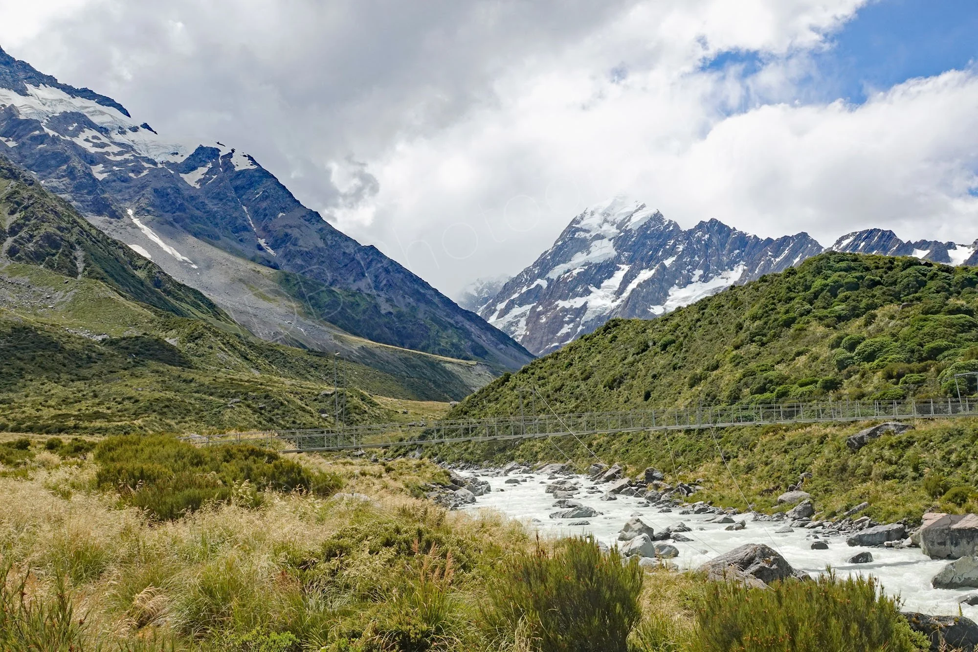 View of River Hooker with swing bridge,  snow capped mountains and clouded sky on the Hooker Valley track,  Canterbury, New Zealand.