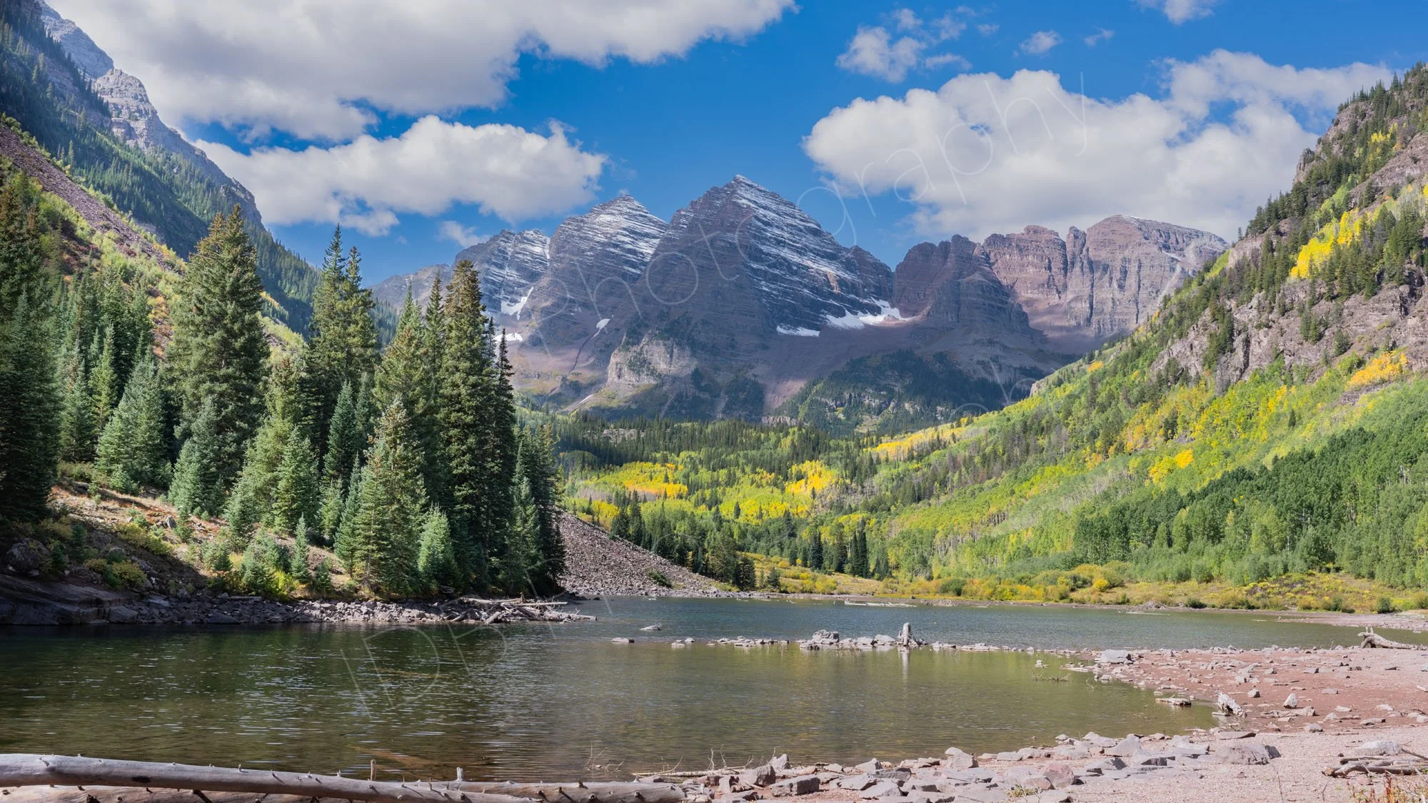 Maroon Bells Lake, Colorado and surrounding mountains with snow residue.  Conifers with green and yellow Aspen trees fill the valley floor; blue sky and clouds. 1.