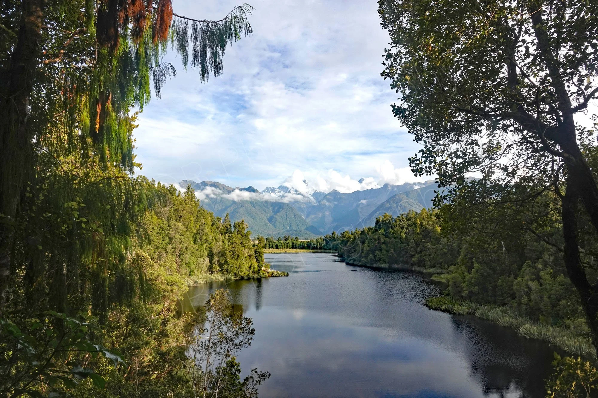 Narrow stretch of Lake Matheson, West Coast, NZ  with cloud reflections, flanked by trees and mountains in the distance.